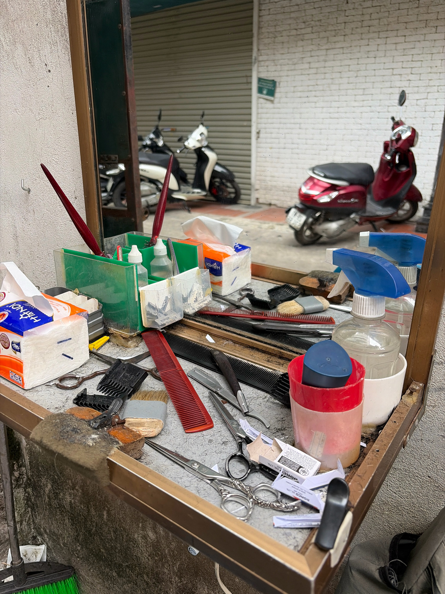 The image depicts a barber's station, likely situated outdoors in Hà Nội, Vietnam, with a reflection in a mirror revealing a street scene. The station itself is cluttered with barbering tools and supplies. In the foreground, on a metallic surface, are various items including scissors of different sizes, combs (both red and black), a razor blade, a shaving brush, and two spray bottles, one with a blue nozzle. There are also several packs of tissues and some small paper boxes. A green plastic organizer holds more brushes and bottles. The background, visible in the mirror's reflection, shows two motorbikes parked on a paved surface, with a white brick wall and a corrugated metal shutter behind them, typical of a storefront in Vietnamese cities. The overall impression is one of a functional, perhaps mobile or roadside, barber setup. There are no people visible in the image.