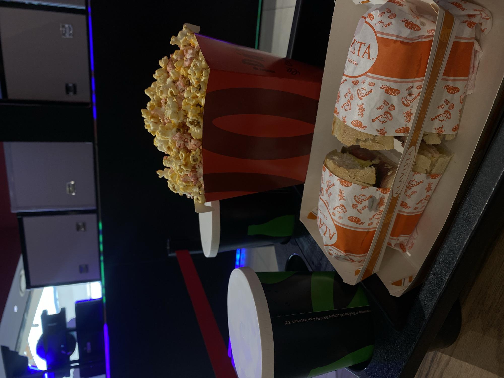 The image displays a collection of movie-style snacks and drinks arranged on a dark surface, likely a table or an armrest with cup holders, against a dark, indoor background illuminated by blurred artificial lights. A large, red and dark-red striped popcorn bucket, overflowing with yellow and pink-tinged popcorn, is prominently positioned on the left.
To the right, two elongated sandwiches or subs are presented within a cardboard tray. Each sandwich is wrapped in white paper featuring orange patterns of bread, vegetables, and fruit. The sandwiches appear partially unwrapped, revealing fillings that include meat and pickles. Below these food items, two dark green or black cups with white lids are visible in cup holders. The background features dark panels with glowing blue and green light lines, suggesting a low-light entertainment environment such as a cinema.
The overall scene depicts an individual or individuals enjoying refreshments, likely during a movie screening or a similar event.
Visible text includes:
* On the sandwich wrappers: "ALA VISTA", "ESSEN" (likely part of DELICATESSEN), "DELICA", and "TA".
* On one of the drink cups: "a registrada de The Coca-Cola Company. D.R. © The Coca-Cola Company. 2025."