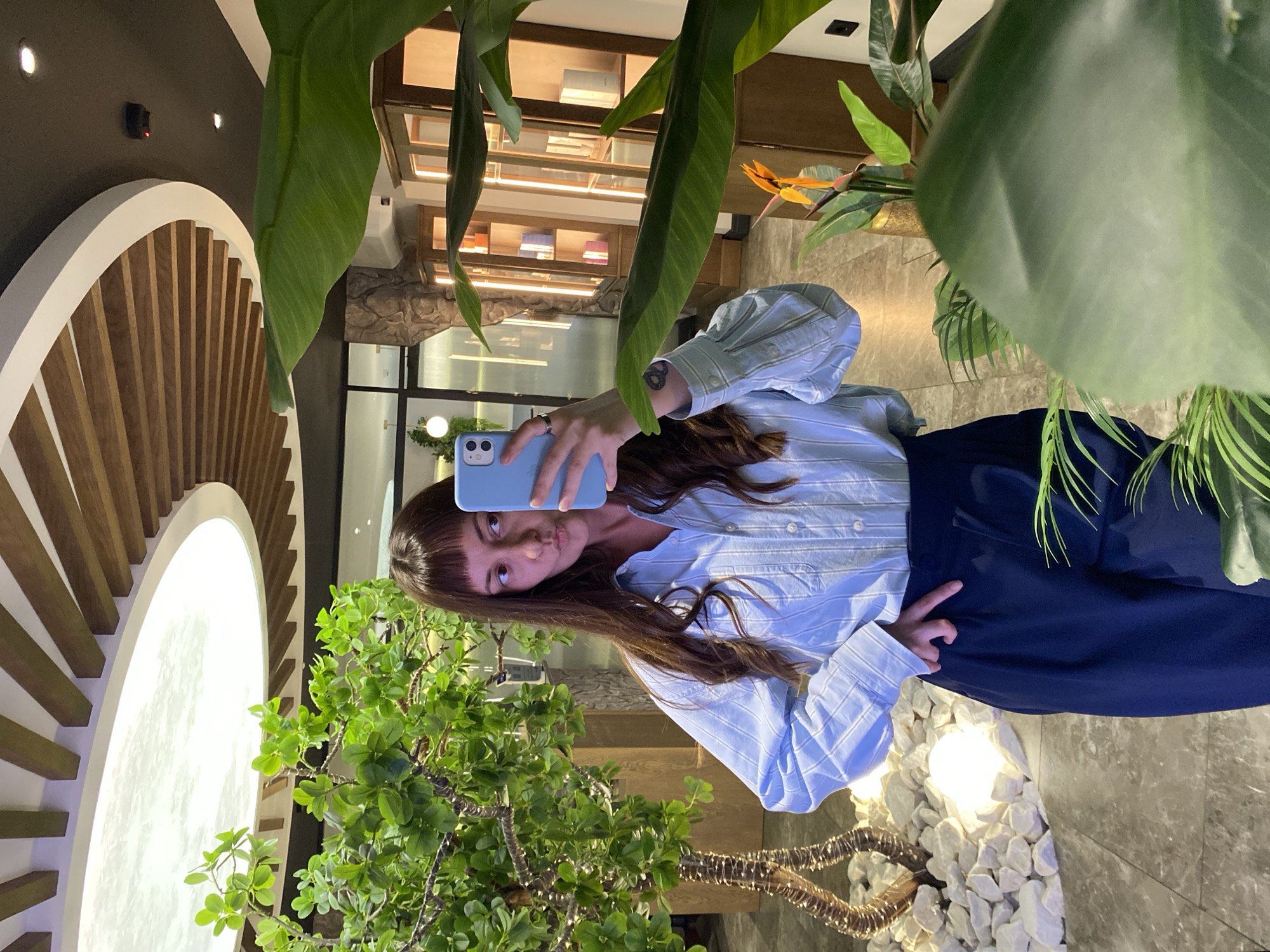 A young woman with brown hair and bangs takes a mirror selfie in a modern, indoor setting. She is wearing a light blue and white striped button-down shirt and dark blue wide-leg pants. Her left hand is on her hip, and she is holding a light blue smartphone in her right hand, angled to capture her reflection.
The background features a variety of elements:
- To the left, there's a decorative tree with bright green foliage, surrounded by white stones and illuminated by a bright white globe light.
- Behind the tree, a textured stone wall is visible.
- To the right, large green leaves of a tropical plant dominate the foreground, obscuring some of the background.
- Further back, wooden cabinets with glass fronts are illuminated, displaying various items.
- Above, the ceiling has a unique design with curved wooden beams and a large circular illuminated panel.
The overall impression is of a stylish, contemporary interior space, possibly a hotel lobby, a cafe, or a well-designed retail establishment in Votkinsk, Russia. The lighting is soft and ambient, creating a warm and inviting atmosphere.