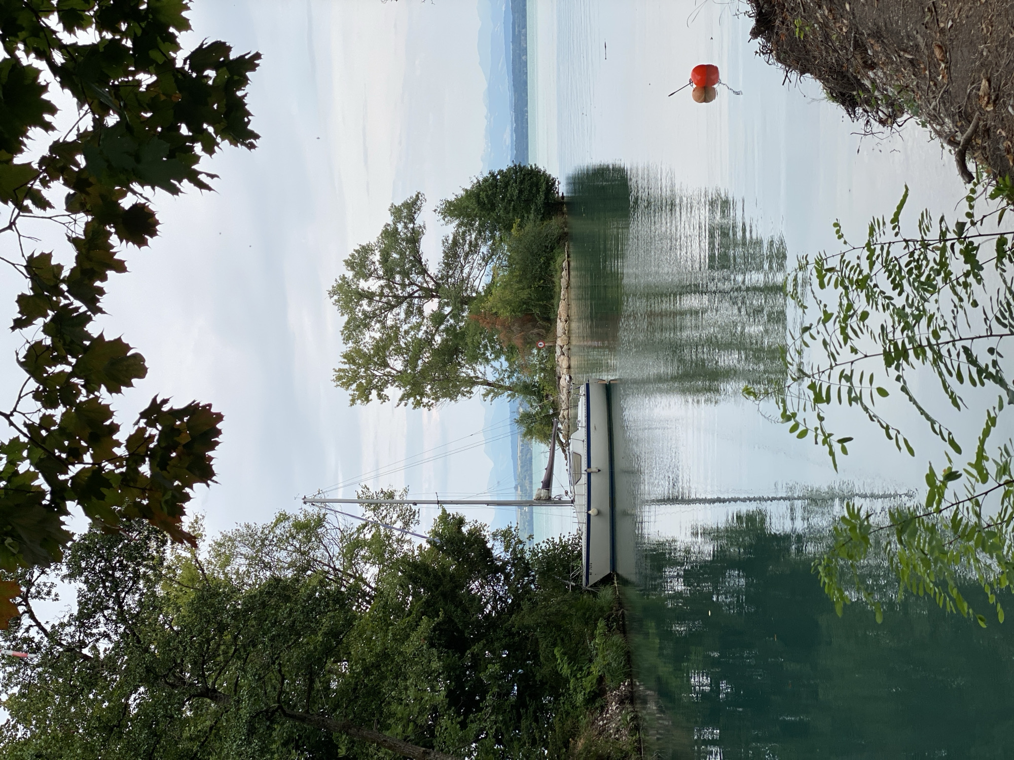 A sailboat is docked in a tranquil, emerald-green canal leading to a serene lake. The sailboat, white with blue trim, has its mast raised and sails furled. Lush green trees line the left bank of the canal, with their reflections mirroring in the still water. In the distance, across the lake, a cluster of islands with trees is visible, and beyond that, hazy blue mountains rise against a pale, overcast sky. A bright orange and red buoy floats on the right side of the lake. The foreground features the edge of the canal and the beginning of the lake, with some vegetation and soil visible on the right bank. The scene appears to be in Cortaillod, Switzerland, likely along the shore of Lake Neuchâtel.
