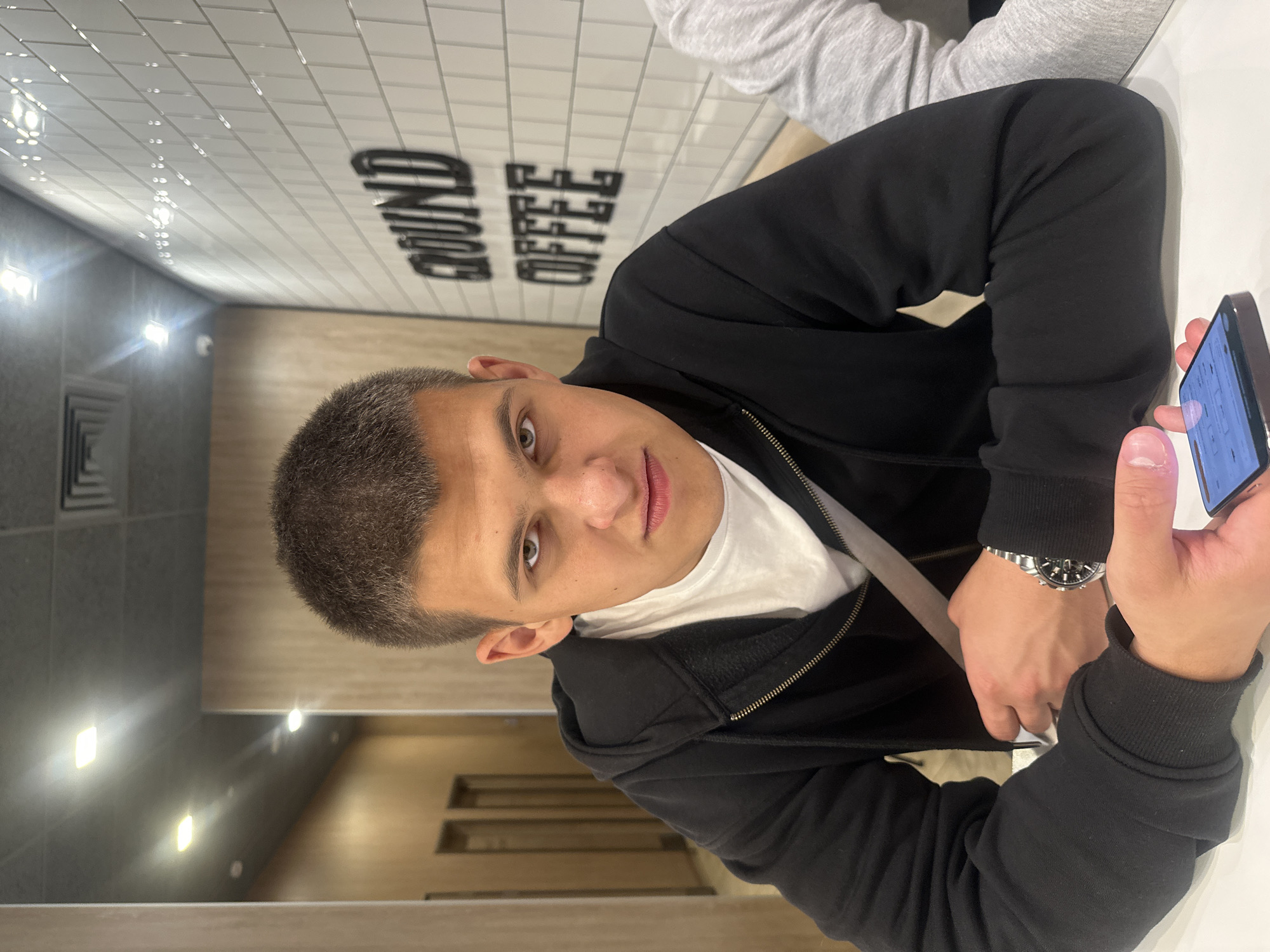 A young man with short brown hair and a serious expression sits at a table in what appears to be a coffee shop. He is wearing a black zip-up hoodie over a white t-shirt and a watch on his left wrist. His left arm is resting on the table, and his right hand is holding a smartphone. The background shows a tiled wall with the words "GROUND COFFEE" in large black lettering and a hallway with wooden doors. The lighting suggests it is indoors. The image does not contain any specific information or visual cues that would allow for its location to be identified as Birobidzhan, Russia.