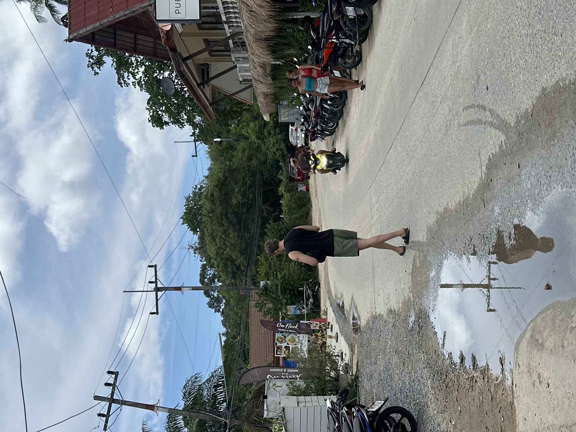 A street scene in Koh Phangan, Thailand, features a road with a light grey asphalt surface. On the left side of the road, a gravel shoulder is lined with parked motorcycles and a white building with a black awning. A large puddle of water reflects the sky and a utility pole on the gravel shoulder. A man in a black tank top and olive green shorts walks away from the camera on the road, carrying a dark object. Further down the road, a yellow scooter with two occupants, a man and a woman, approaches. On the right side of the road, a building with a red tiled roof and a thatched awning is visible. Several motorcycles are parked in front of this building. Two women, one in a blue tank top and white shorts, the other in a red bikini top and white shorts, walk towards the camera along the sidewalk in front of the building. Trees and utility poles line the street, and the sky is partly cloudy.