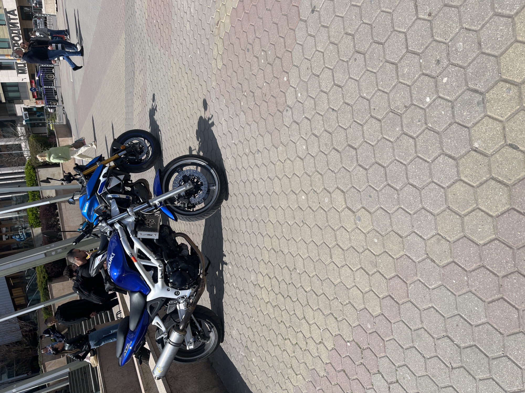 The image depicts a city street in Sofia, Bulgaria. The foreground is dominated by two motorcycles, both blue and white Suzuki SV models, parked perpendicular to the curb. Several individuals are standing near the motorcycles, appearing to be engaged in conversation and inspection of the vehicles. The background shows a building with signage in Cyrillic script, and pedestrians walking along the sidewalk. The street is paved with a mix of gray and red-toned hexagonal pavers. The weather appears sunny, casting shadows of the motorcycles and people on the pavement.