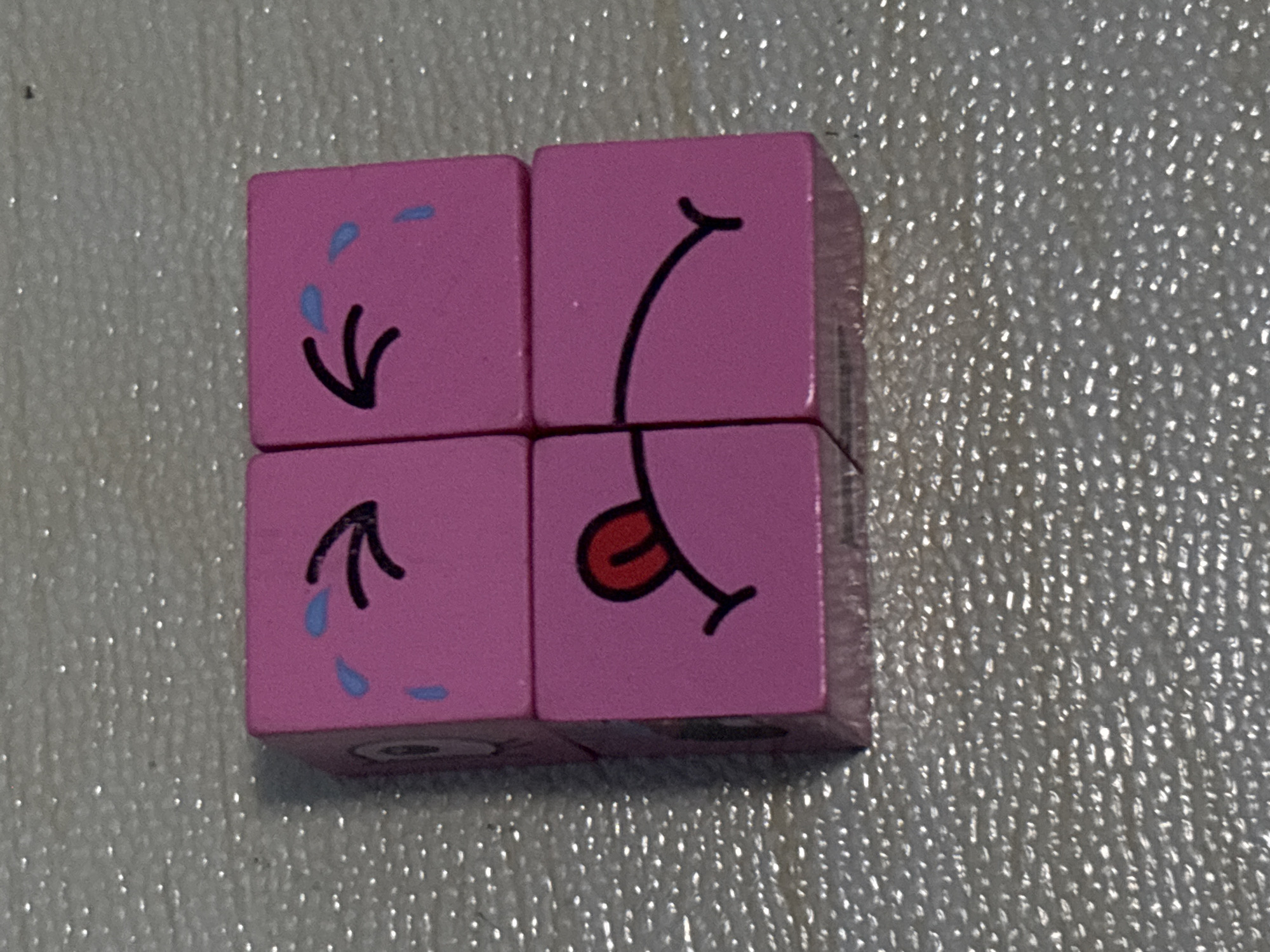 The image shows a close-up, overhead view of four pink toy blocks arranged in a 2x2 grid on a textured, speckled gray surface. The blocks are the same size and appear to be made of wood or plastic. Each block displays a portion of a cartoon face:
The top-left block shows a closed eye with a blue tear and a curved black line representing an eyebrow or part of a nose.
The top-right block shows a closed eye with a blue tear and a curved black line representing an eyebrow or part of a nose.
The bottom-left block shows a red tongue sticking out with a black outline, and a portion of a mouth.
The bottom-right block shows a curved black line that appears to be the lower part of a mouth.
Together, the blocks form a face with crying eyes and a mouth with a tongue sticking out, suggesting an expression of sadness or being overwhelmed. The background surface is highly reflective with numerous small, bright specks.
No location in Bekasi, Indonesia, or any people are visible in this image.