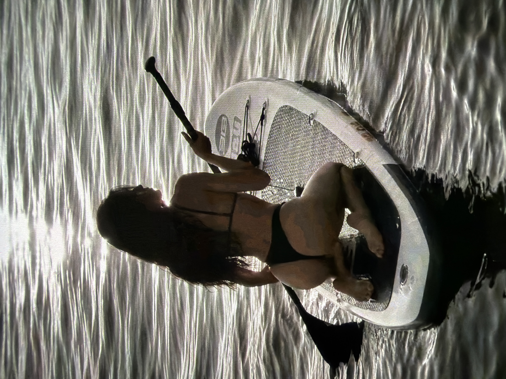 A woman is paddleboarding on a body of water. She is wearing a black bikini and is kneeling on the paddleboard, holding a paddle. The paddleboard is white and has a black traction pad. The water is choppy and reflects the sunlight. The scene is set in Brunswick, Germany.