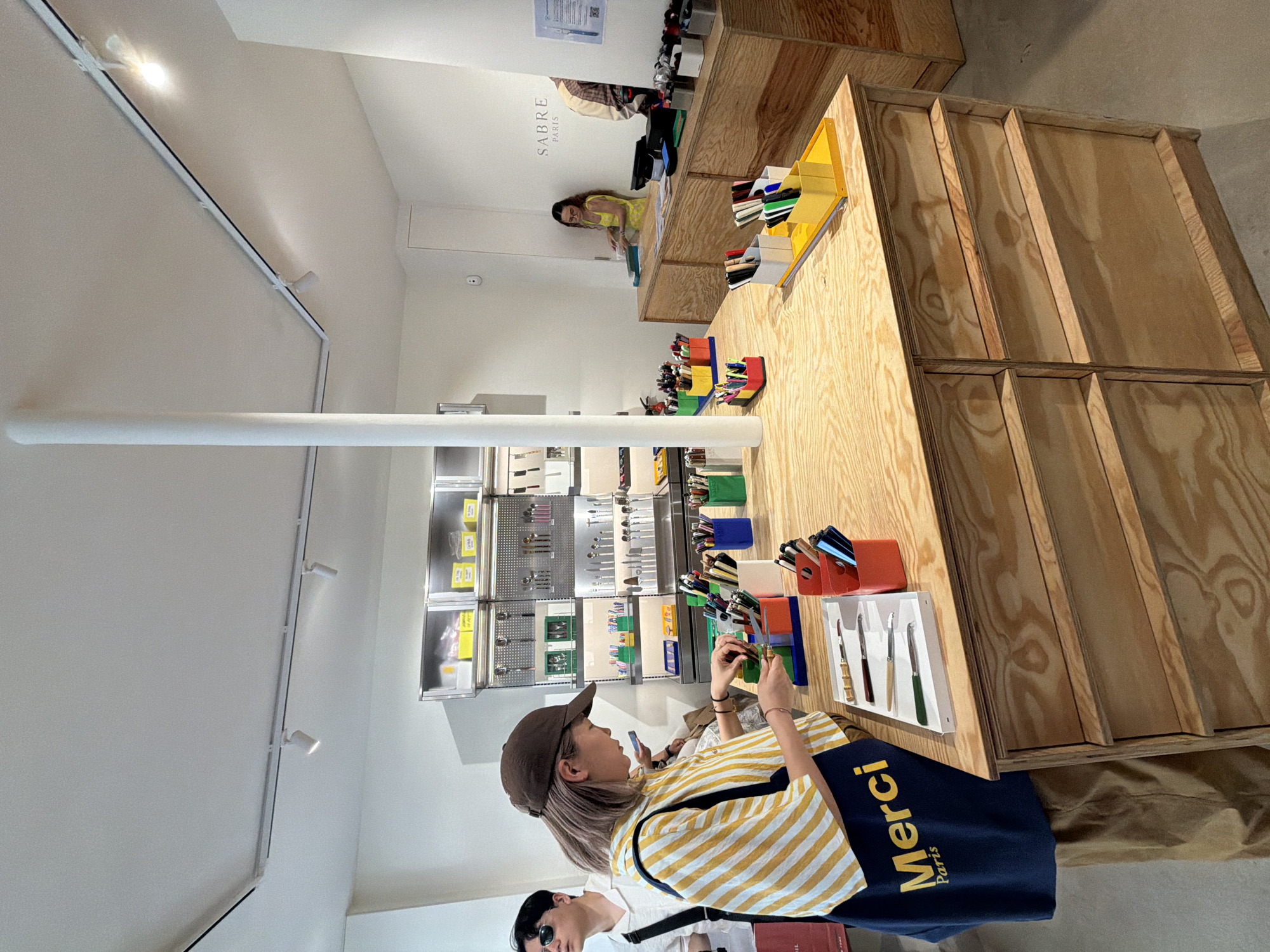 The image depicts the interior of a retail space, likely a shop, located in Paris, France. A woman wearing a brown cap and a yellow and white striped shirt is looking at items displayed on a wooden countertop. She is holding a product in her hands and inspecting it. A dark blue shopping bag with the word "Merci Paris" printed on it is visible near her. Behind her, there is another person partially visible, wearing a white shirt and sunglasses. The shop's interior features display shelves stocked with various items, including cutlery. A sign with the text "Sabre Paris" is visible in the background. The interior design includes a white ceiling with track lighting and a large, white support beam. The countertop is made of light-colored wood with a noticeable grain pattern and has a stepped design. The overall lighting in the shop is bright, creating a clean and modern atmosphere.