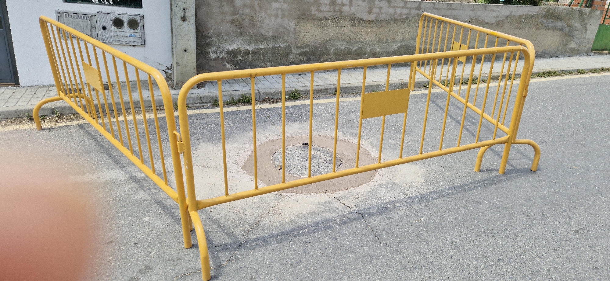 In an outdoor urban setting, a yellow metal barrier is placed on asphalt, partially blocking access to a manhole cover. The barrier is comprised of horizontal bars and vertical supports, with two blank rectangular yellow signs attached to the upper bar. The manhole cover, visible beneath the barrier, has a circular pattern and is surrounded by a circular area of disturbed earth and sand. The ground surrounding the manhole cover appears to be wet or recently worked. To the left of the barrier, a white building wall with two utility boxes is visible, one with a gray door and the other with circular, glass-like elements. The building wall is adjacent to a paved sidewalk with a yellow line marking. Beyond the sidewalk, sparse green vegetation grows. The background consists of a textured, gray concrete wall and a further section of a building with a green gate. The overall scene suggests ongoing or completed construction or maintenance work in a street environment.