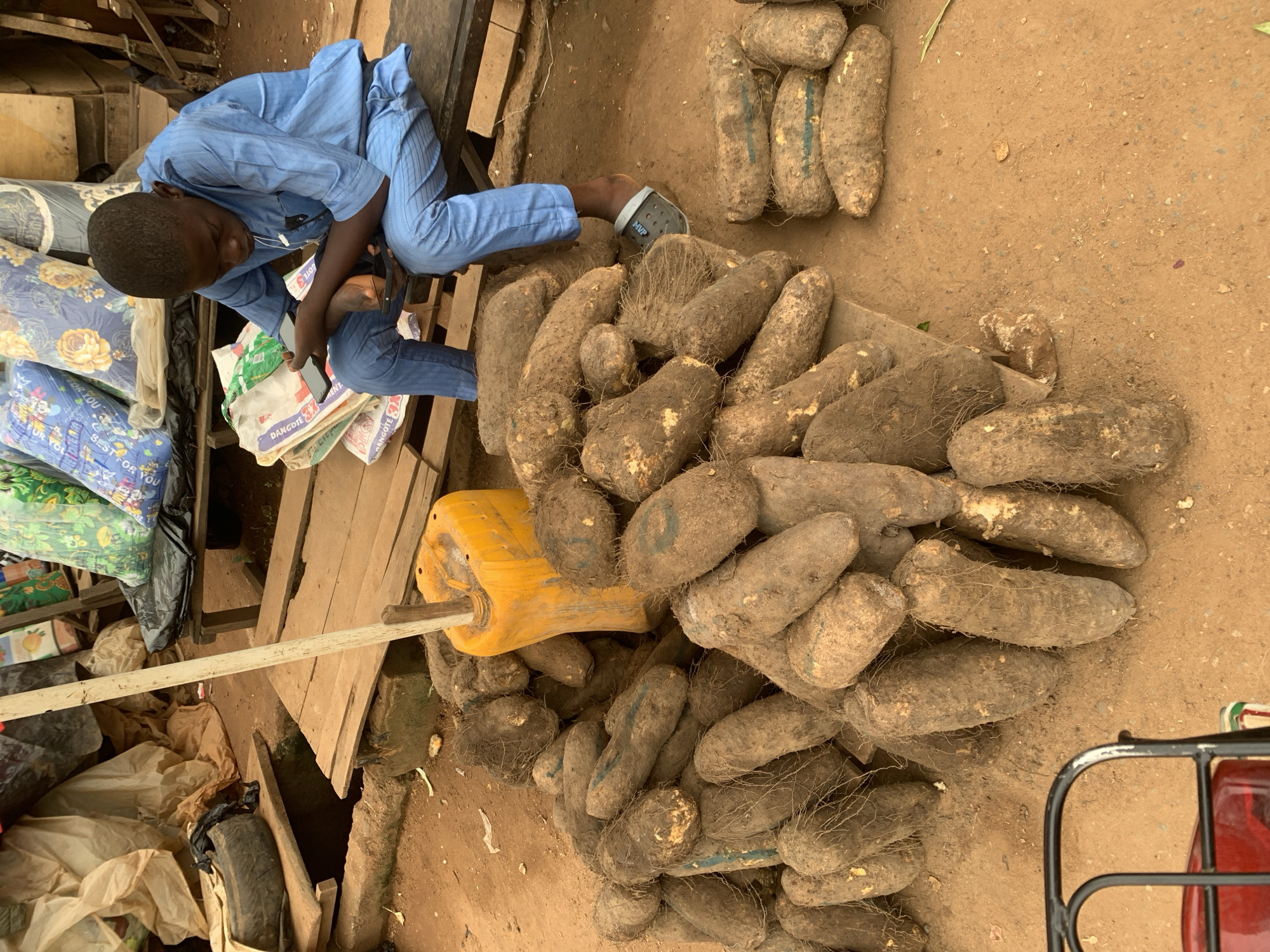 In Shagamu, Nigeria, an outdoor market scene unfolds as a young boy, dressed in a light blue traditional outfit, sits on a wooden bench engrossed in his mobile phone. He is surrounded by large piles of yams, a staple food, which are displayed on the dusty ground and on wooden planks. To his left, a yellow jerrycan is propped upright by a wooden pole. In the background, various goods are stacked, including what appear to be mattresses and sacks, suggesting a bustling commercial environment. The ground is a mixture of dirt and sand, typical of an open-air market in the region.