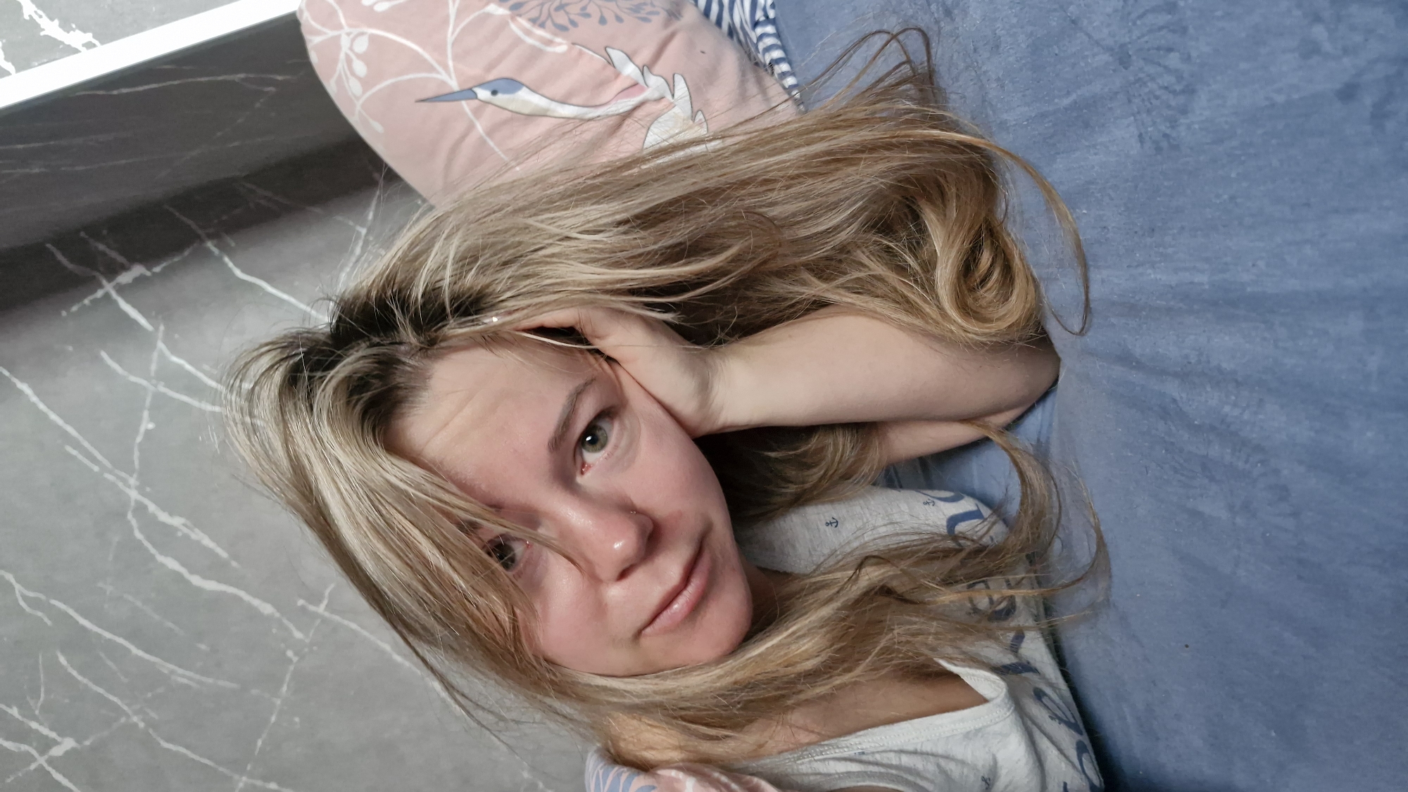 A woman with long, light brown and blonde hair is lying down, with her head resting on her hand. She appears to be indoors, with a gray, marble-patterned surface to her left, possibly a countertop or wall. A pink pillow with bird designs is visible behind her head, and she is lying on a blue surface, likely a bed or sofa. She has fair skin and light-colored eyes and is wearing a white top with a blue anchor pattern. The focus is on her face and upper body. There is no definitive information about Otchor, Russia within the image.