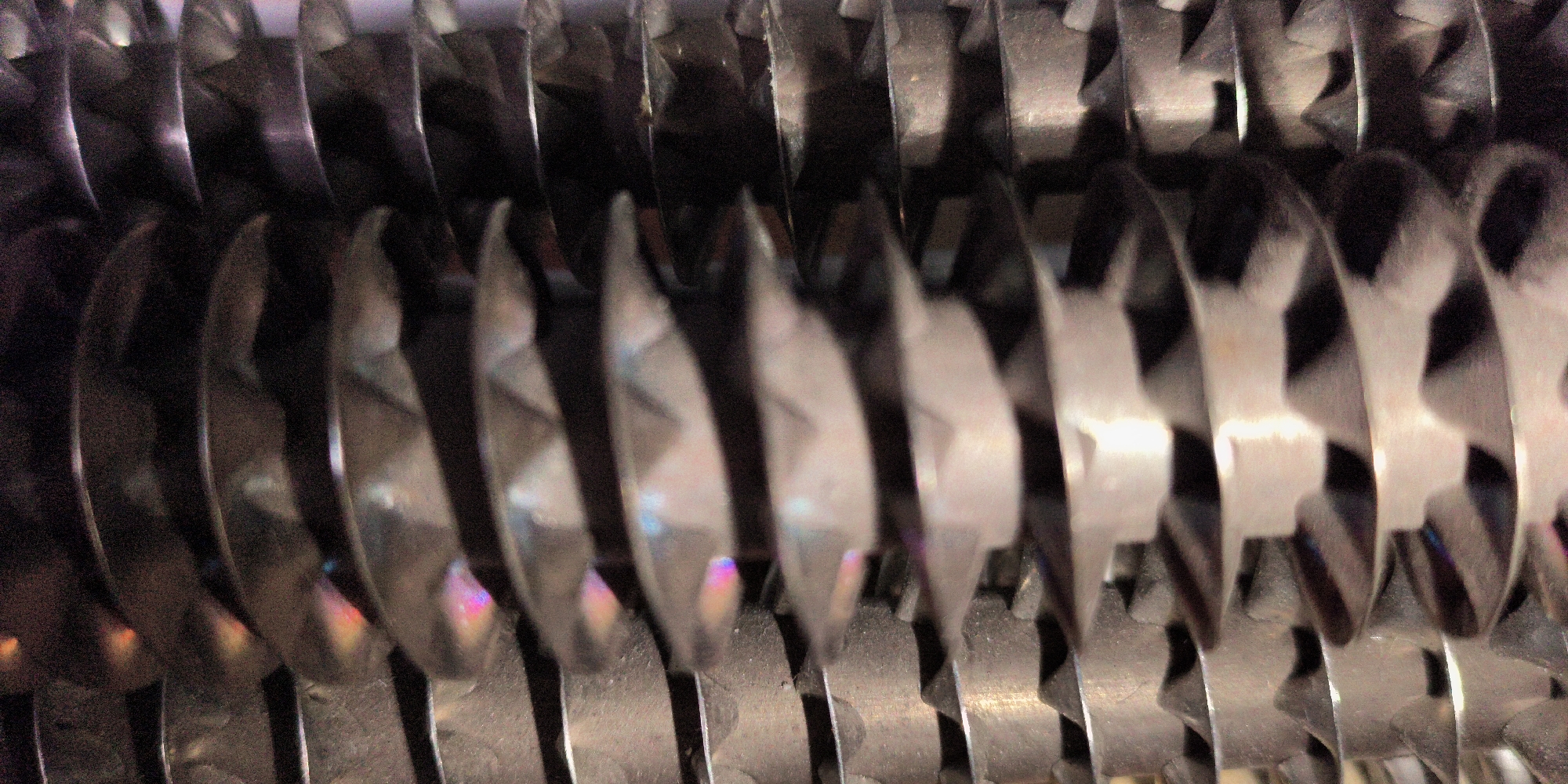 The image shows a close-up, abstract view of a series of metal blades, likely from a shredder or a similar industrial machine. The blades are arranged in a spiral or helical pattern, with sharp edges visible. They are metallic, with some areas reflecting light and showing iridescent colors, possibly due to oil or residue. The background is dark and out of focus, emphasizing the texture and form of the blades. There are no people, animals, or any identifiable environmental features. The image does not contain any information about a specific city or location.