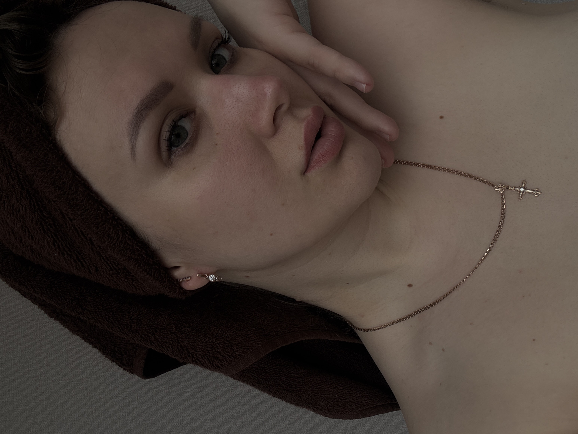 A person is shown in a close-up, tilted portrait view. The individual has fair skin, light eyes, and pink lips. Their hair is covered with a brown towel. They are wearing a delicate gold chain necklace with a cross pendant and a small earring. The person's hand is raised near their face. The background appears to be a neutral grey surface. The location is unspecified and cannot be inferred from the image.