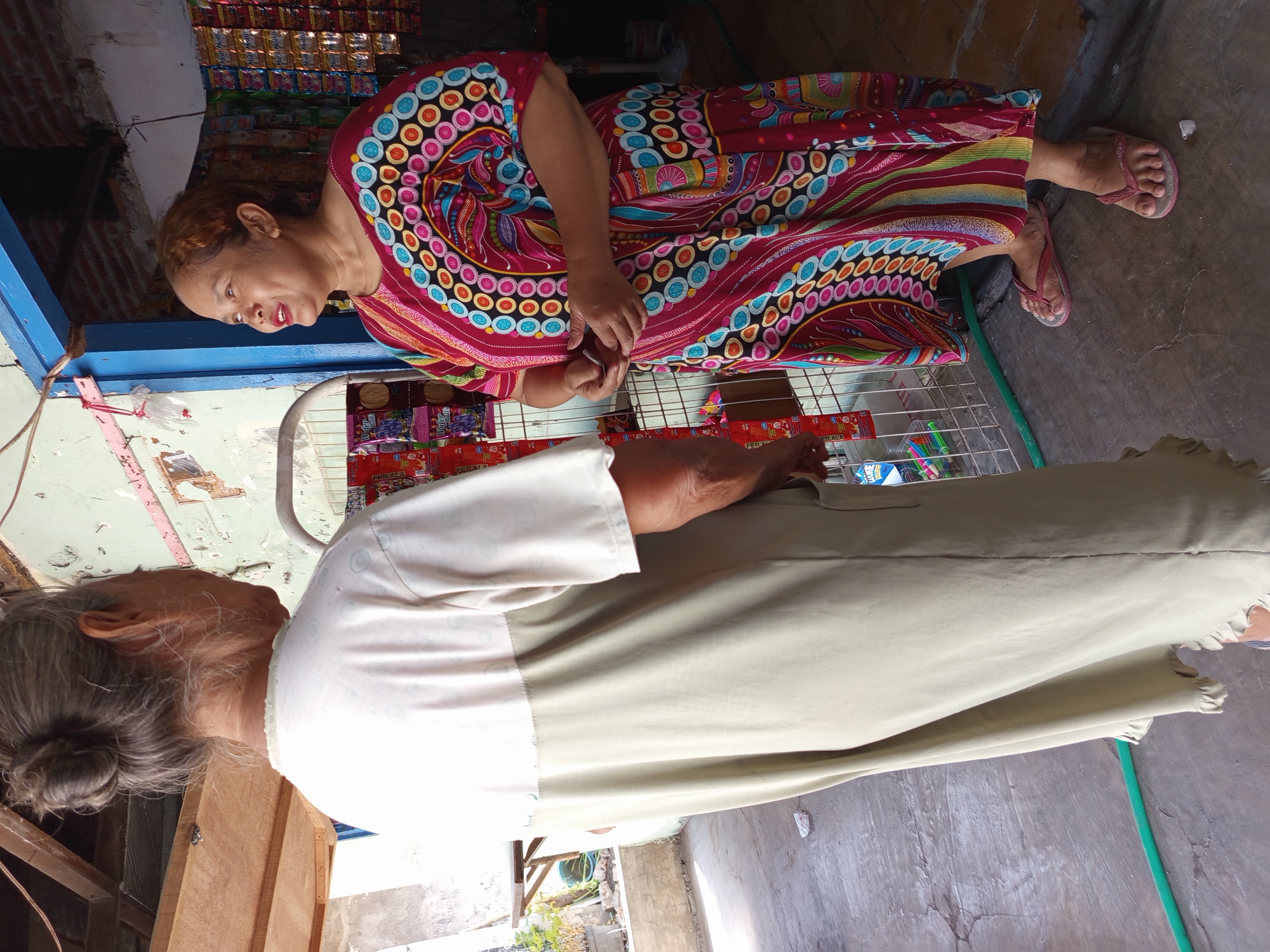 Two women stand on a concrete ground in front of a small store in Tebel, Indonesia. The woman on the left, seen from behind, wears a light beige dress with a white top section. Her hair is tied up in a bun. The woman on the right faces the woman on the left, looking upwards and to her left. She is wearing a brightly colored dress with a circular and stripe pattern in red, pink, yellow, blue, and black. She has brown hair and is wearing pink flip-flops. They are standing near a wire rack displaying various packaged goods. The store entrance is visible behind the woman on the right, showing more packaged goods on shelves. The environment appears to be outdoors, with some daylight filtering in.