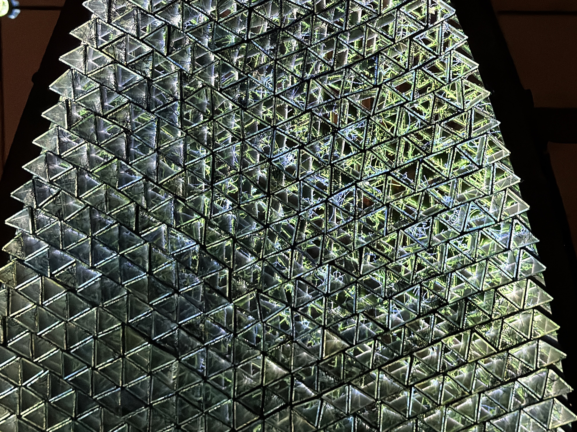 The image displays a close-up, abstract view of a textured surface composed of numerous small, transparent triangular prisms arranged in a repeating pattern. These prisms are stacked and layered, creating a complex, geometric three-dimensional effect. Light passes through and reflects off the facets of the prisms, resulting in a shimmering and prismatic appearance.
The overall color palette is dominated by cool tones of grey and blue, with hints of green and yellow visible within the prisms, suggesting reflections of an environment or internal lighting. The background is dark, with some areas of shadow and light, hinting at an indoor setting or a controlled lighting environment. The texture appears somewhat rough or fibrous in places, visible through the transparent prisms, adding another layer of detail to the composition.
There are no discernible people, animals, or recognizable objects in the foreground or background. The focus is entirely on the abstract interplay of light, shadow, and geometric form created by the arrangement of the triangular prisms. The image evokes a sense of intricate design and optical complexity, perhaps hinting at architectural elements, a decorative screen, or a scientific model. There is no indication of the time of day, weather, or specific emotions. No text is visible in the image.