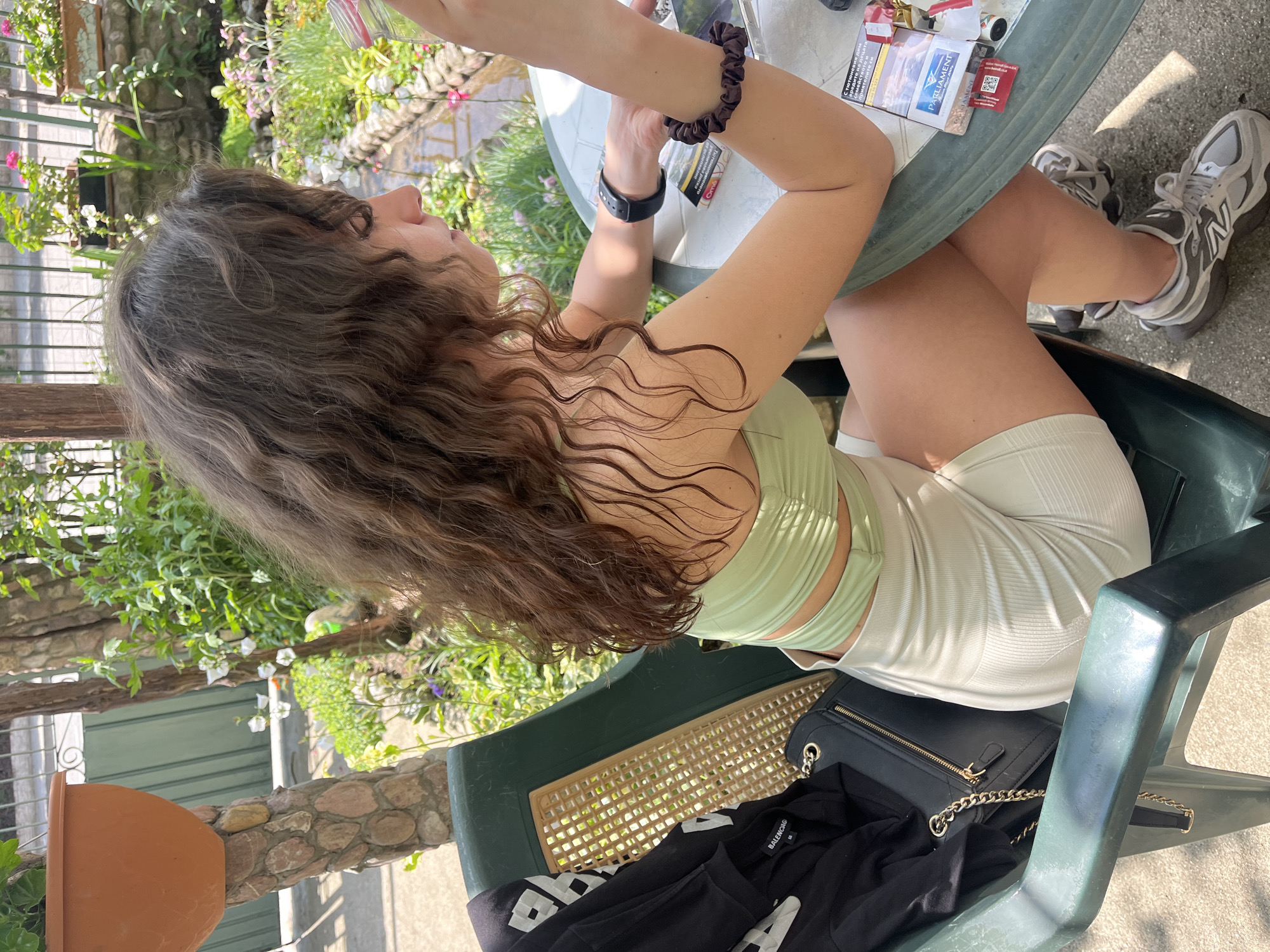 A young woman with long, wavy brown hair, seen from a high angle and side profile, is seated outdoors in a green plastic chair. Her head is tilted slightly upwards, and her right arm is raised, with a black scrunchie on her wrist and a black watch. She is wearing a light green, ribbed crop top and light-colored ribbed shorts. Her feet, clad in gray and white New Balance sneakers, are visible.
In front of her, on a green round table, rests a pack of Parliament cigarettes and a small red item, possibly a label or QR code. A black handbag with a gold chain strap is placed on an adjacent green chair, along with a black garment that has white text, likely "BALENCIAGA."
The setting is an outdoor patio or garden area, characterized by lush greenery, various flowering plants, and stone elements in the background. A green slatted fence or gate and a building with green shutters are also partially visible. The scene is brightly lit by the sun, casting strong shadows, indicating daytime. Visible text includes "Parliament" on the cigarette pack, "BALENCIAGA" on the black garment, and the "N" logo of New Balance on her shoes. Some Cyrillic text is also present on the cigarette pack's warning label.