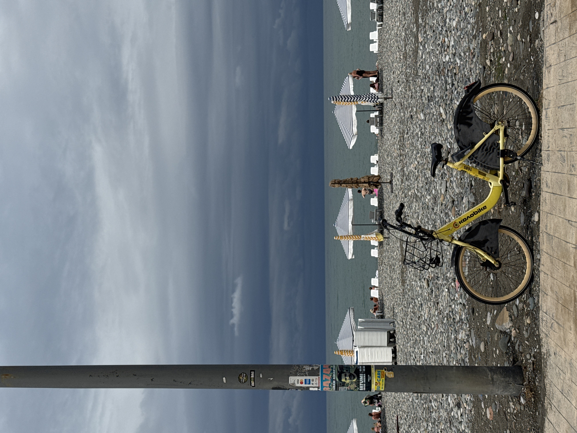 This is a photograph taken in Batumi, Georgia, on a cloudy day. The foreground features a yellow rental bicycle parked on a pebble beach. The bicycle has a basket on the front and a kickstand. To the left of the bicycle is a lamppost with several stickers and posters attached, one of which advertises "Batumi Circus Arena". In the background, the sea is visible under a dark, overcast sky. There are beach chairs and umbrellas set up on the pebble shore, with a few people scattered around, some sitting on chairs and others standing near the water.