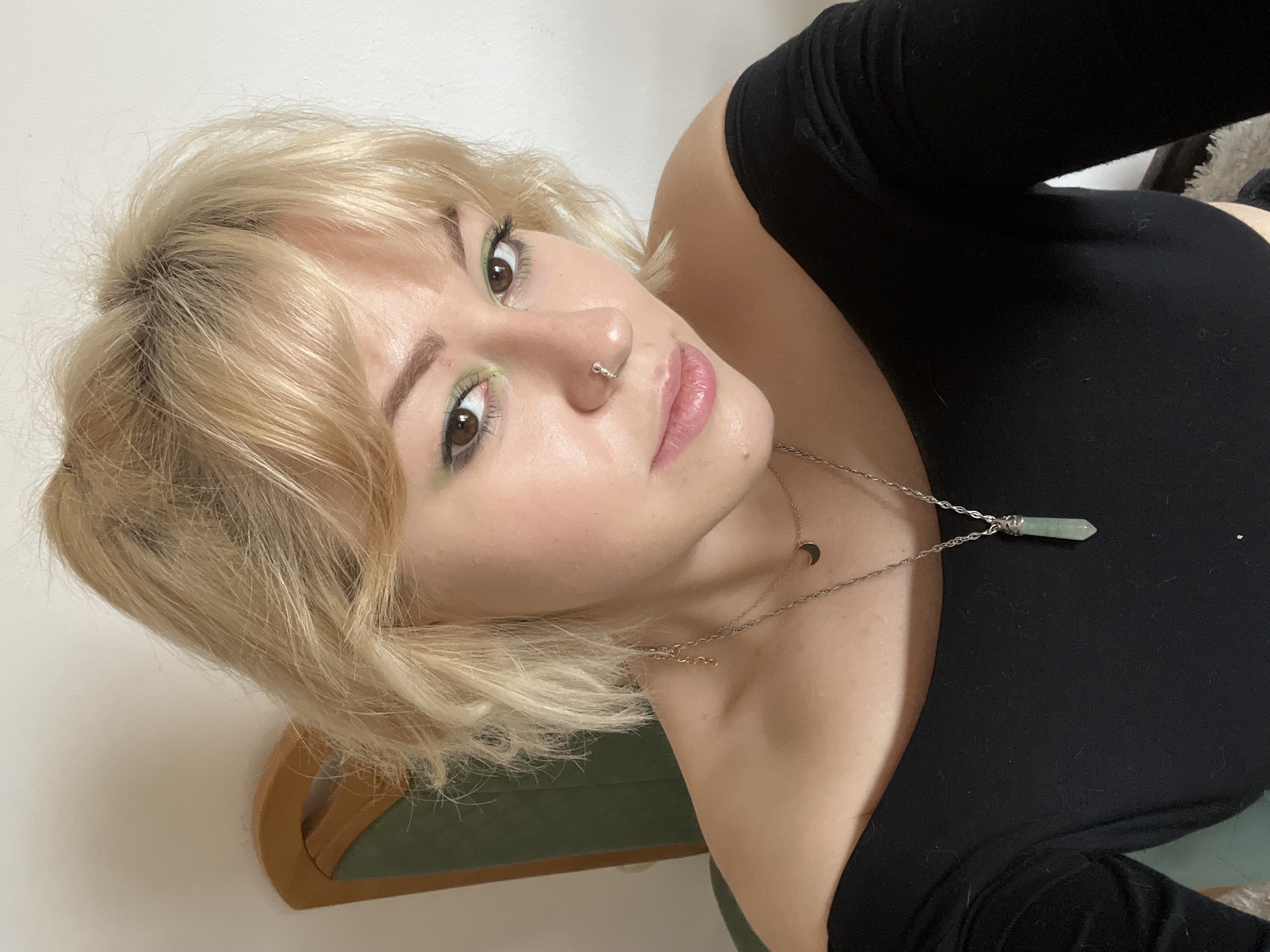 A close-up, high-angle studio shot depicts a young woman with blonde hair styled in a choppy bob with bangs. She has light skin, brown eyes with green eyeshadow, and a nose ring in her left nostril. She is wearing a black, form-fitting long-sleeved top. Two necklaces are visible around her neck: one with a small moon pendant and the other with a green crystal pendant. She is seated in a chair with a green upholstered back, set against a plain white wall. The photo provides no information to suggest this image was taken in Budva, Montenegro.