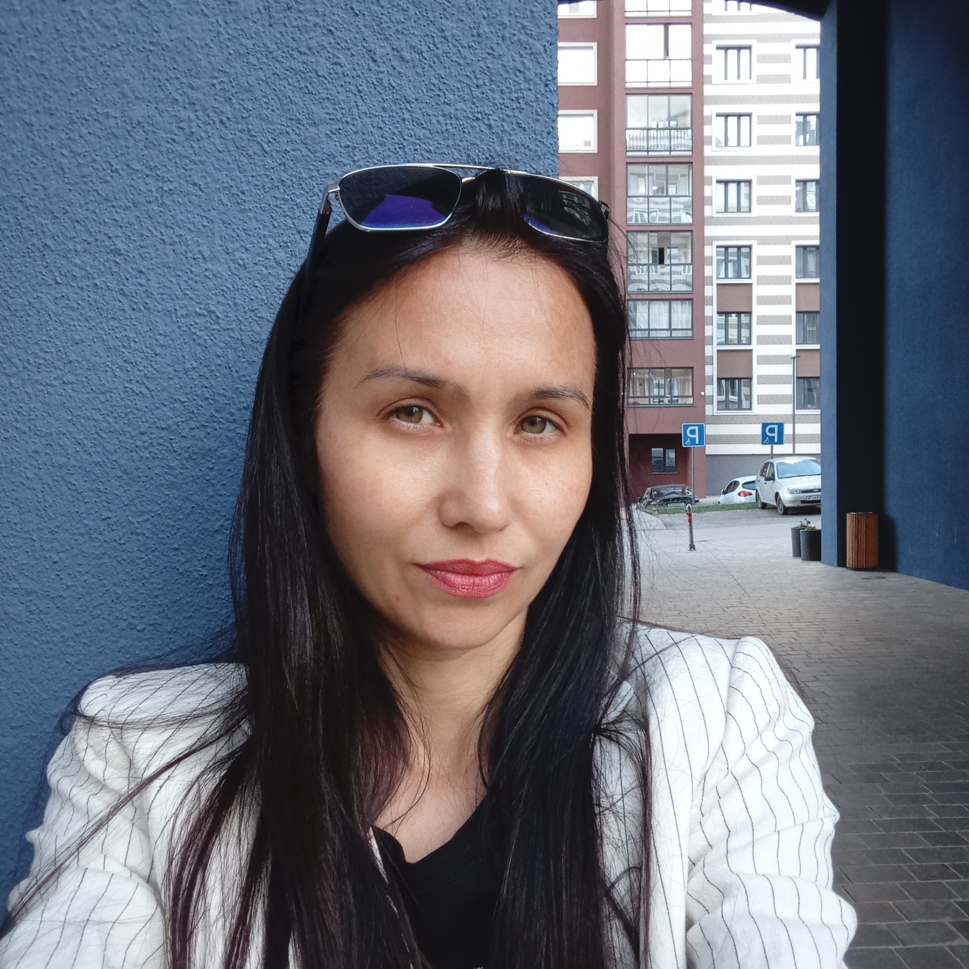 A woman is captured in a head-and-shoulders portrait, facing directly towards the camera. She has long, dark hair, light greenish-brown eyes, and is wearing red lipstick. Sunglasses with reflective blue lenses are resting on top of her head. Her attire includes a white blazer with thin black pinstripes over a dark top.
She is positioned against a textured, matte blue wall, which appears to be part of an architectural feature like a building's entrance or archway. Beyond her and to her right, an urban residential scene unfolds. The background features modern multi-story apartment buildings with varied facades, including one with distinctive brown and white horizontal stripes. The ground is paved with grey interlocking bricks, and a street or courtyard is visible further back.
In the mid-ground, two cars (one white, one dark) are parked near two blue rectangular signs, each displaying a white 'P' and an arrow, indicating parking. There are also potted plants and a cylindrical trash bin with a textured, possibly wooden, exterior. The scene is illuminated by natural daylight, suggesting it is daytime with diffused light, possibly due to an overcast sky or the time of day. The woman holds a neutral and composed expression. The location context is Pervomaysky, Russia.