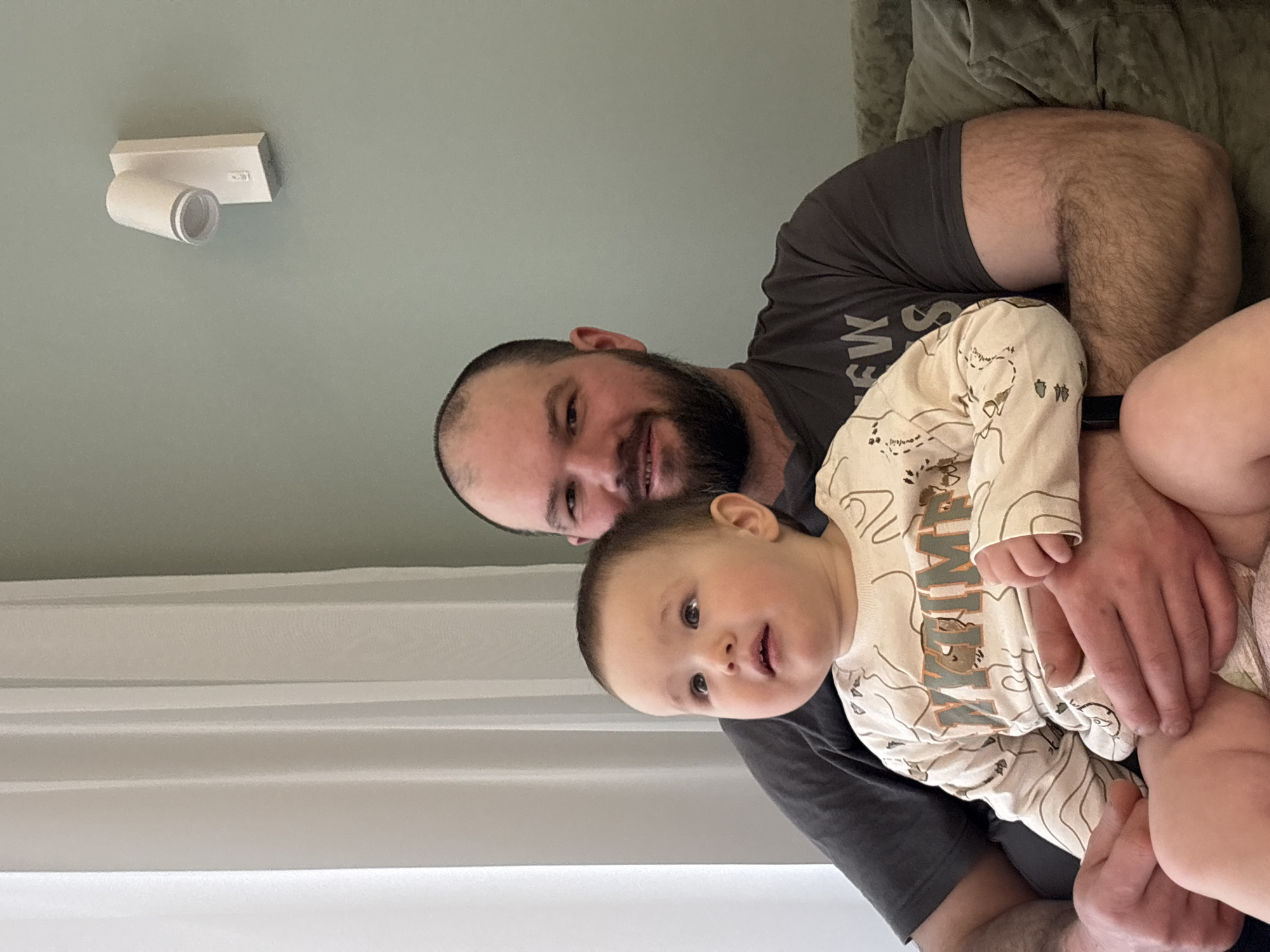 A man with a beard and short hair is holding a baby. The baby is wearing a patterned, light-colored onesie. The man is wearing a dark gray t-shirt. They are indoors. A white spotlight fixture is visible on the ceiling. A curtain or blind is in the background. There is no indication of Izhevsk, Russia in the image.