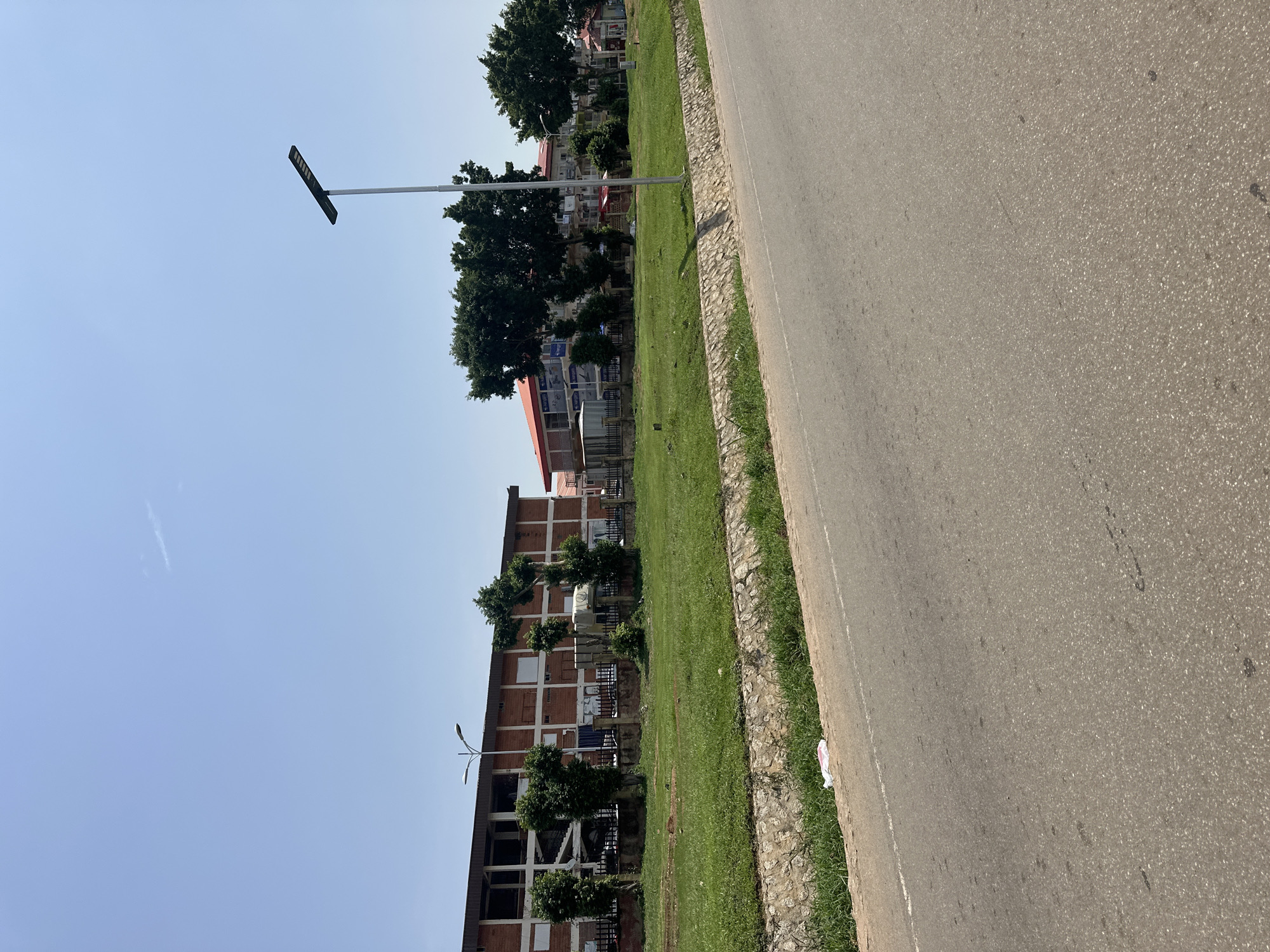 A wide, exterior, eye-level, horizontal shot shows a paved road in the foreground, with a grassy median and a multi-story brick building with open-air floors behind it. A tall, modern street lamp stands in the grassy median, to the right of the building. The building appears to be an office building or residential complex, with several trees planted in front of it. The sky is clear and blue. The scene appears to be a street in Abuja, Nigeria.