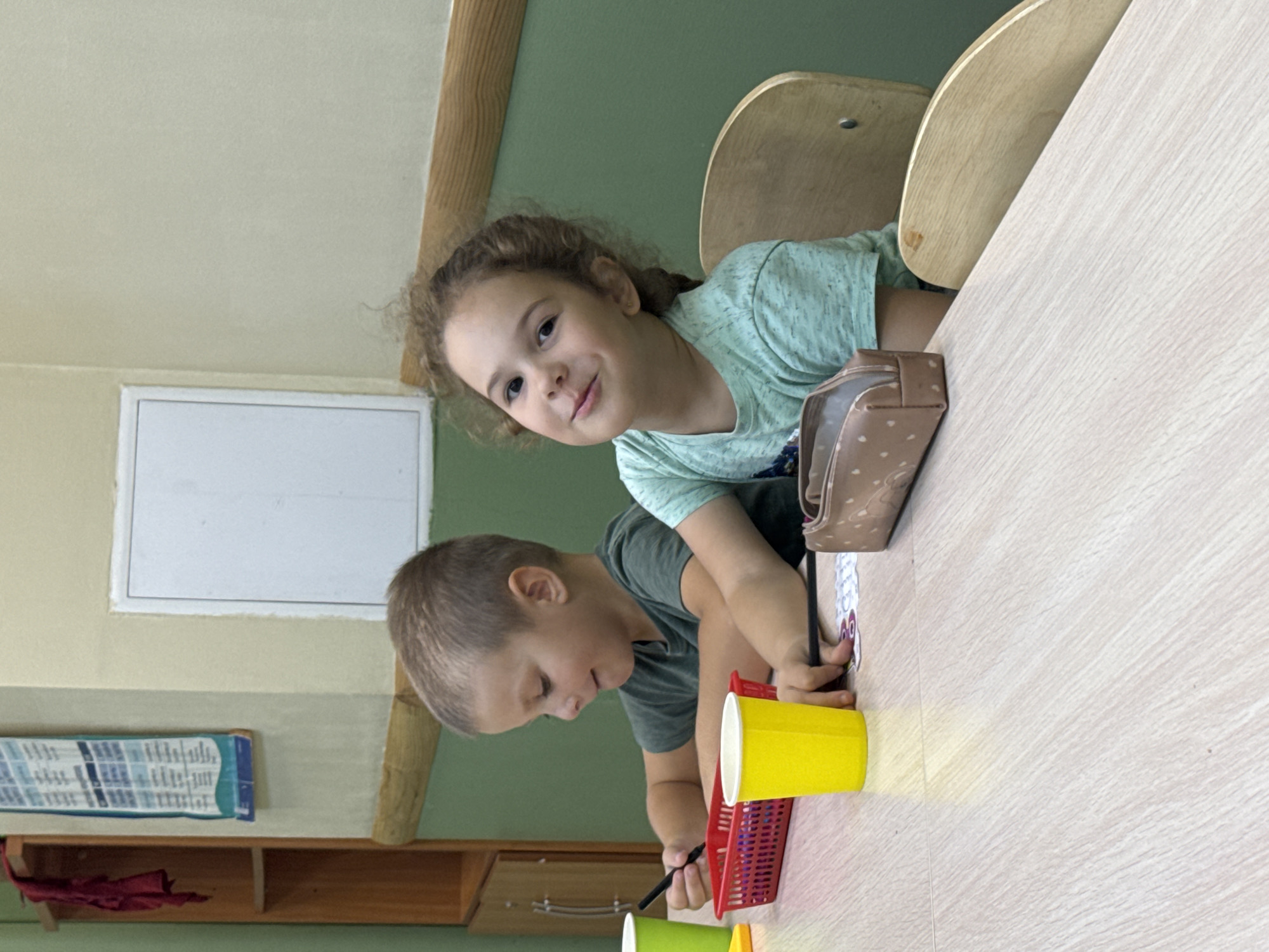 Two young children, a boy and a girl, are seated at a light-colored wooden table in what appears to be a classroom or activity room. The boy, on the left, has short brown hair and is wearing a dark green t-shirt. He is holding a black marker and looking down at a red basket filled with colorful items, possibly art supplies. In front of him is a bright yellow paper cup. The girl, on the right, has curly brown hair and is wearing a light teal t-shirt. She is looking directly at the camera with a slight smile. In front of her is a small, light brown pencil case with a subtle pattern. The background shows a light green wall with a wooden trim near the bottom and a white rectangular fixture on the upper part of the wall. To the left, there is a wooden shelf unit with a red piece of fabric hanging from the top shelf. A blue and white poster is also visible on the wall near the shelves. The overall lighting suggests an indoor environment, possibly during daylight hours. The scene implies a moment of artistic activity or a break during a lesson. The location is identified as a classroom in the city of Perm, Russia.