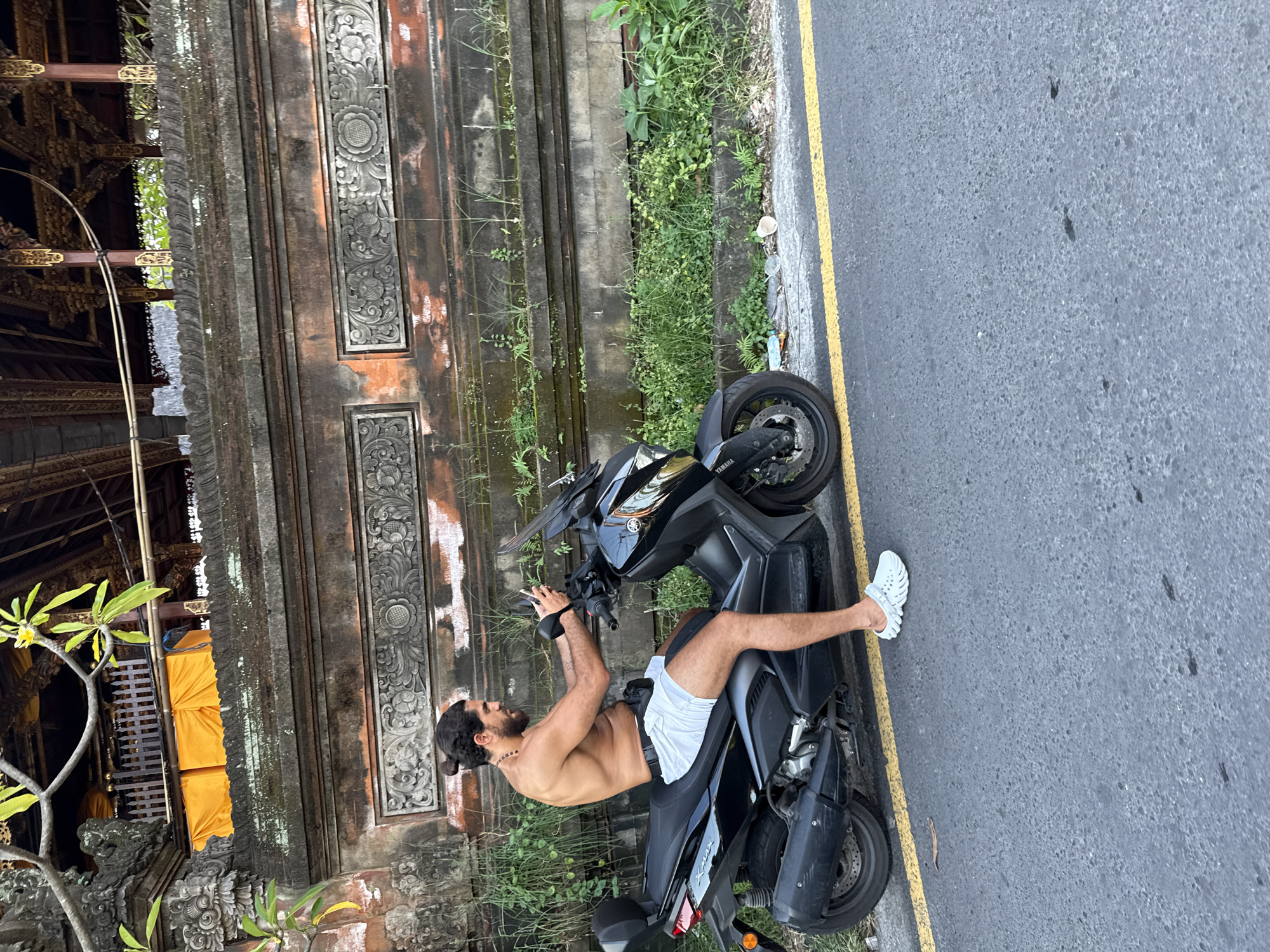 An eye-level, medium outdoor shot features a shirtless man with a beard reclining on a black Yamaha Xmax 300 motorcycle, parked on the side of a paved road in North Kuta, Indonesia. The man is wearing white shorts and white shoes. The motorcycle is parked near a yellow road marking. A temple wall with intricate carvings and vegetation stands in the background. Additional plants and vegetation grow along the base of the wall. The weather appears to be overcast.