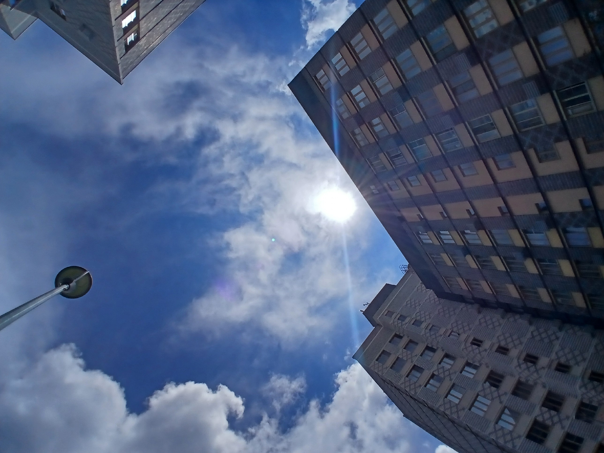 This is an exterior, low-angle, wide shot of two residential buildings in Yekaterinburg, Russia, under a bright, partly cloudy blue sky. The sun is visible, creating lens flare effects. In the foreground, a tall, gray, concrete apartment building with a diamond-shaped patterned facade is on the left. Adjacent to it, and slightly to the right, is another, darker apartment building with yellow and dark gray paneling. Both buildings have numerous windows. In the upper portion of the image, a streetlamp extends into the frame from the top, with its light fixture visible. Another building, part of which is visible in the upper right corner, has a dark, angular facade with geometric window cutouts. There are no people visible in the image.