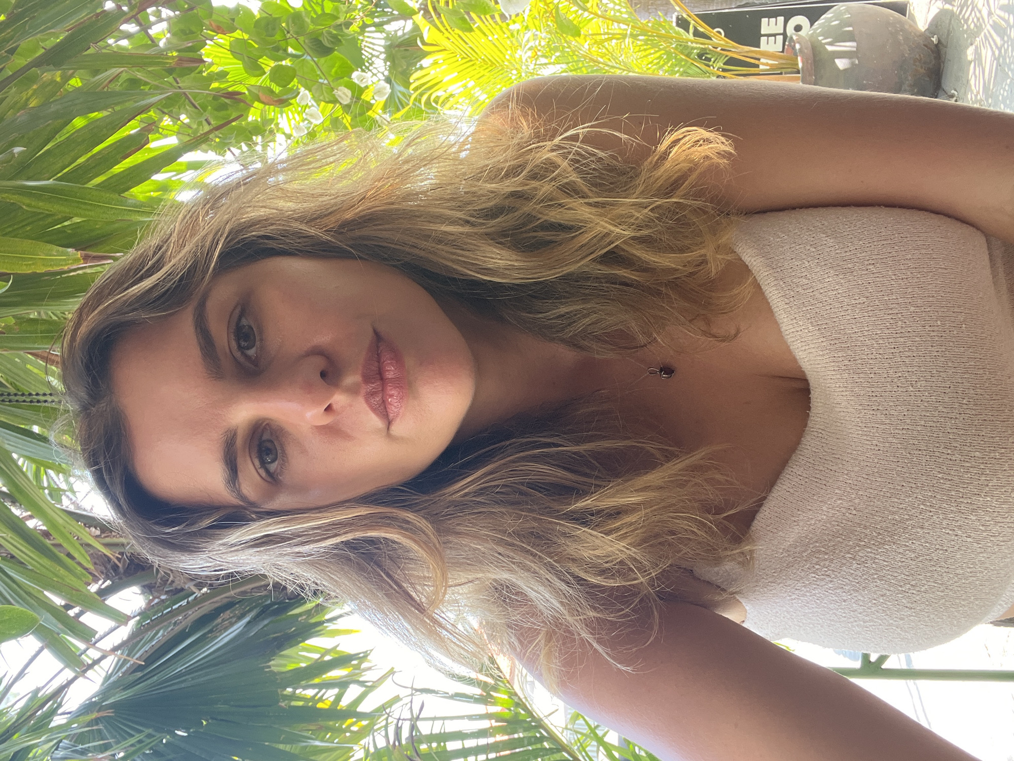 This is a close-up, waist-up, selfie-style portrait of a woman with blonde, wavy hair and light skin, taken outdoors in Mengwi, Indonesia. She is looking directly at the camera with a neutral expression. She is wearing a light-colored, ribbed, strapless top and a simple necklace with a dark pendant.
The background features lush, green tropical foliage, including palm fronds and other leafy plants, bathed in bright sunlight. The lighting creates a warm, dappled effect. To the right, a portion of a brown ceramic pot containing a plant is visible, and a dark sign with partial white text, possibly related to a cafe or establishment, is also present. The overall setting suggests a relaxed, outdoor environment, possibly a cafe patio or garden area within Mengwi.