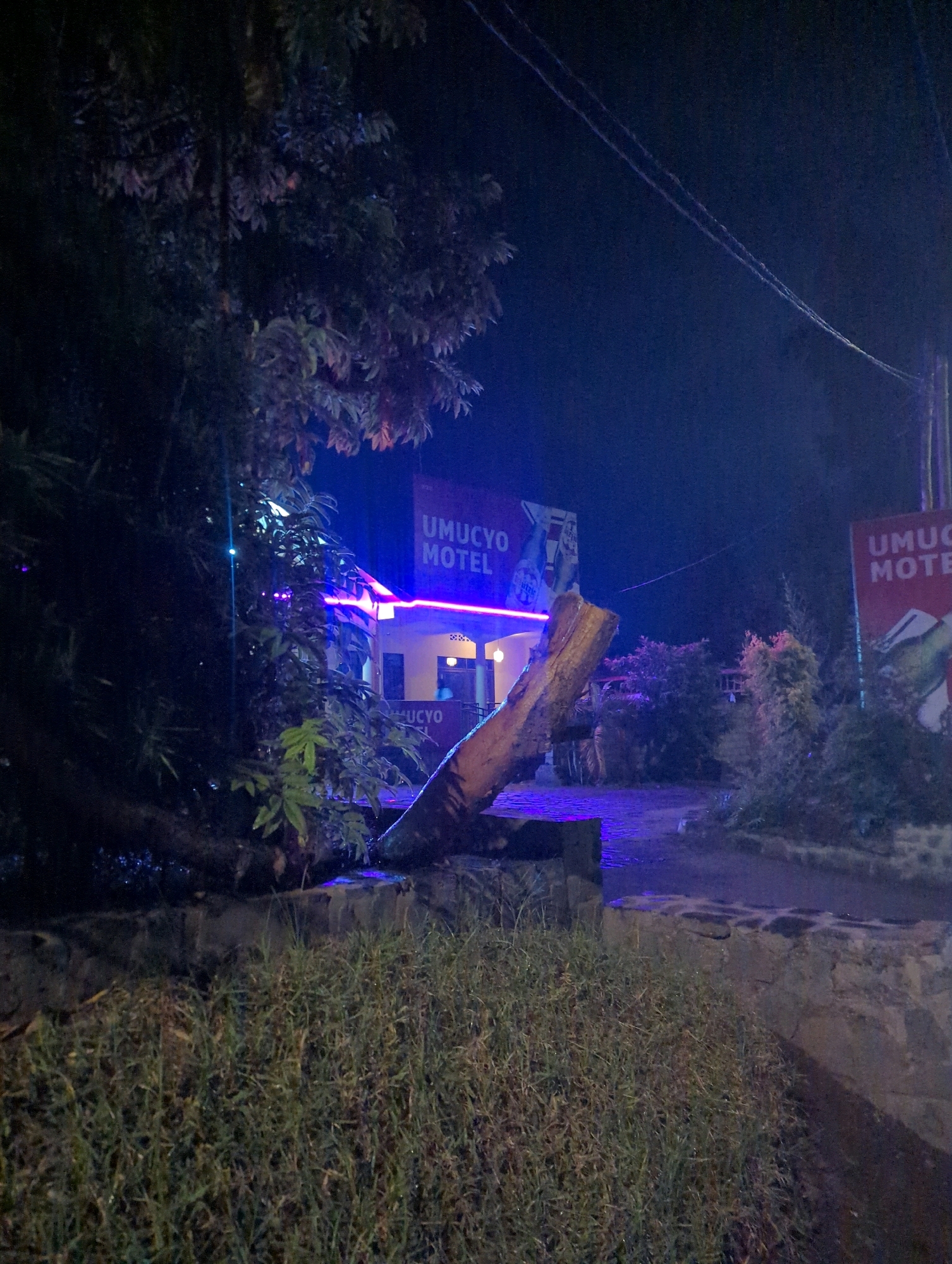 At night, a large tree trunk lies horizontally across a low stone wall in the foreground. Beyond the wall, the grounds of a motel are visible. A sign reading "UMUCYO MOTEL" is illuminated with neon purple lights, as is the building's facade. Another similar sign is visible to the right. The scene is dark, with electric lights providing the primary illumination. The environment appears to be an outdoor area of a motel in Musanze District, Rwanda.