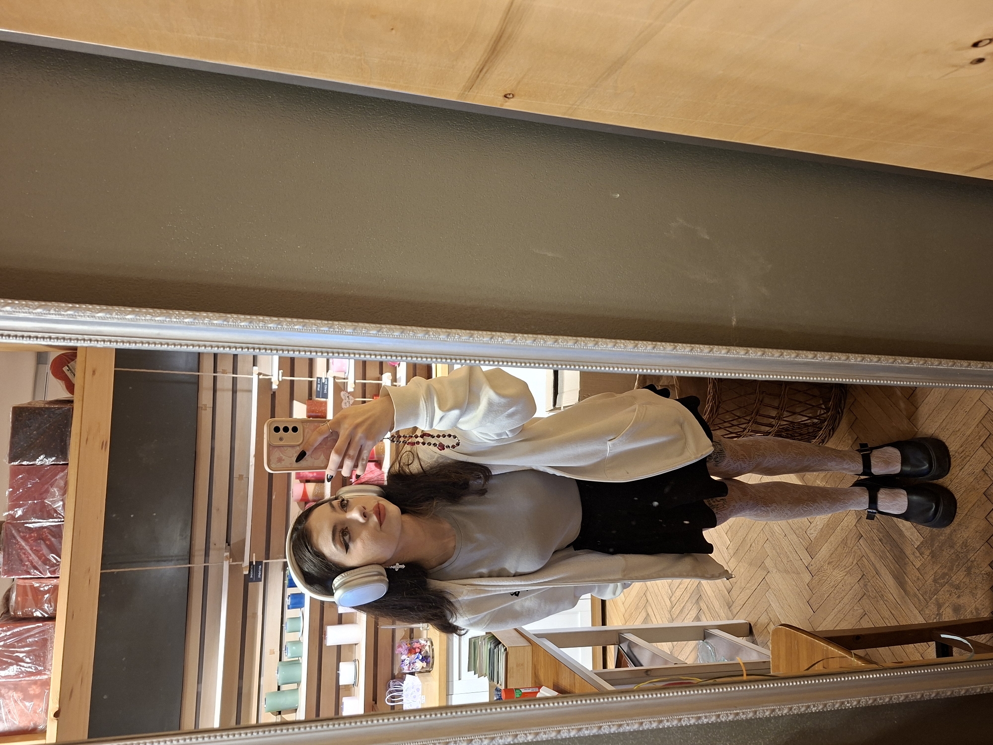 The image is a reflection in a silver-framed mirror. The reflection depicts a person wearing a light-gray shirt, black skirt, white cardigan, white patterned tights, and black Mary Jane shoes. The person is holding a smartphone in their right hand, as if taking a selfie. They are also wearing white headphones.
The reflection also shows a retail environment with shelving displaying various products. The background includes a wooden floor with a herringbone pattern. The ceiling above the mirror appears to be painted gray, with a light wood-colored surface above that.
The location appears to be a shop in Sofia, Bulgaria. The presence of specific items and their display suggests this is a retail setting, possibly a gift shop or boutique.