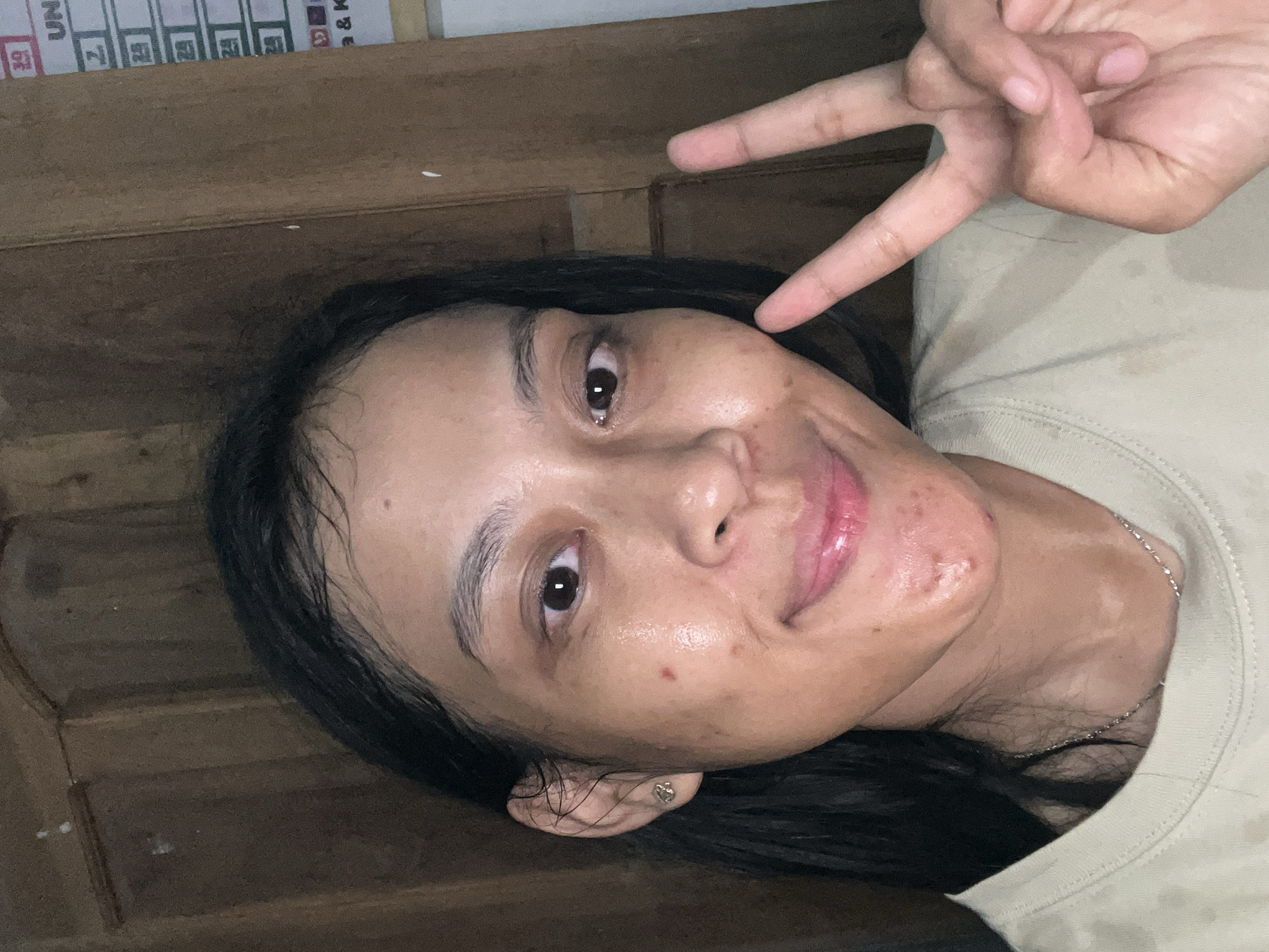 In Kecamatan Pati, Indonesia, a young woman with dark hair and light skin poses for a selfie against a wooden door. She is wearing a light-colored t-shirt and a delicate silver necklace. Her face is turned slightly towards the camera, and she offers a gentle smile with her lips slightly parted, revealing glossy lipstick. She has a small earring in her left ear and several blemishes on her cheeks and chin. Her right hand is raised, forming a peace sign with her index and middle fingers extended. The lighting is dim, casting soft shadows across her features and the textured wooden background. The image appears to have been taken indoors, likely in a residential setting.