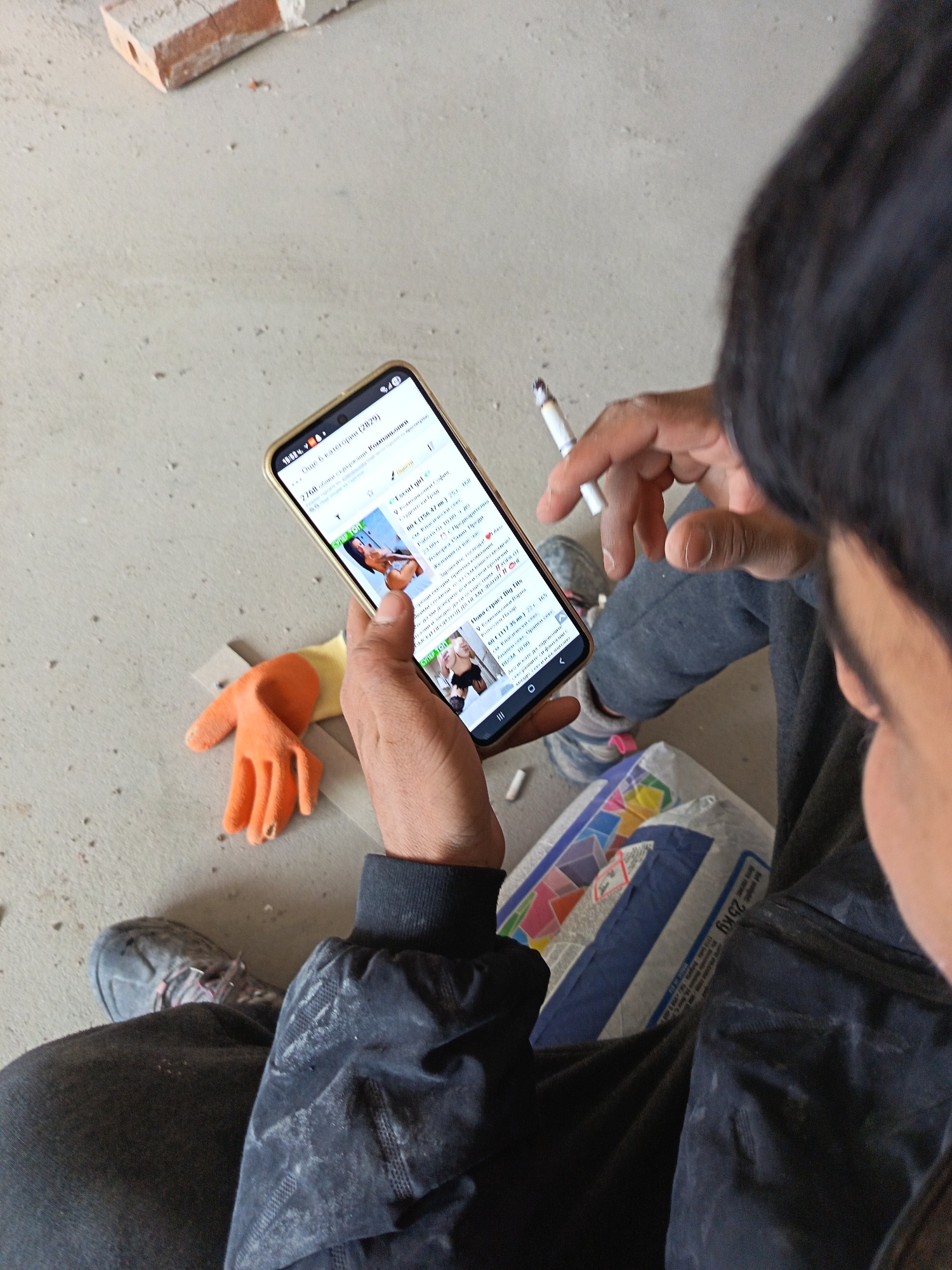 A high-angle, close-up image depicts a person, partially visible from the chest upwards, sitting or squatting on a dusty concrete floor in what appears to be a construction or renovation site in Sofia, Bulgaria. The individual, wearing dark, soiled clothing and worn, dusty sneakers, holds a gold-framed smartphone in their left hand, actively interacting with the screen. The phone displays a webpage with content predominantly in Bulgarian Cyrillic, showing "10:52" as the time and indicating "2760 обяви" (2760 ads) across "6 категории" (6 categories). Specific text on the screen includes "ескорт София" (escort Sofia) and "София" (Sofia), indicating the content's geographical focus. The person's right hand holds a lit cigarette between the thumb and forefinger. Resting on the concrete floor to the left of the phone is a single orange work glove. In the upper left corner, a reddish-brown brick is partially visible. In the lower right corner, a white/grey bag or sack, possibly containing construction material, is visible, marked with colorful geometric patterns, the weight "25 KG," and also bearing the word "София" (Sofia). The environment is characterized by the unpolished, dusty concrete floor and construction-related items, reinforcing the setting within the city of Sofia.