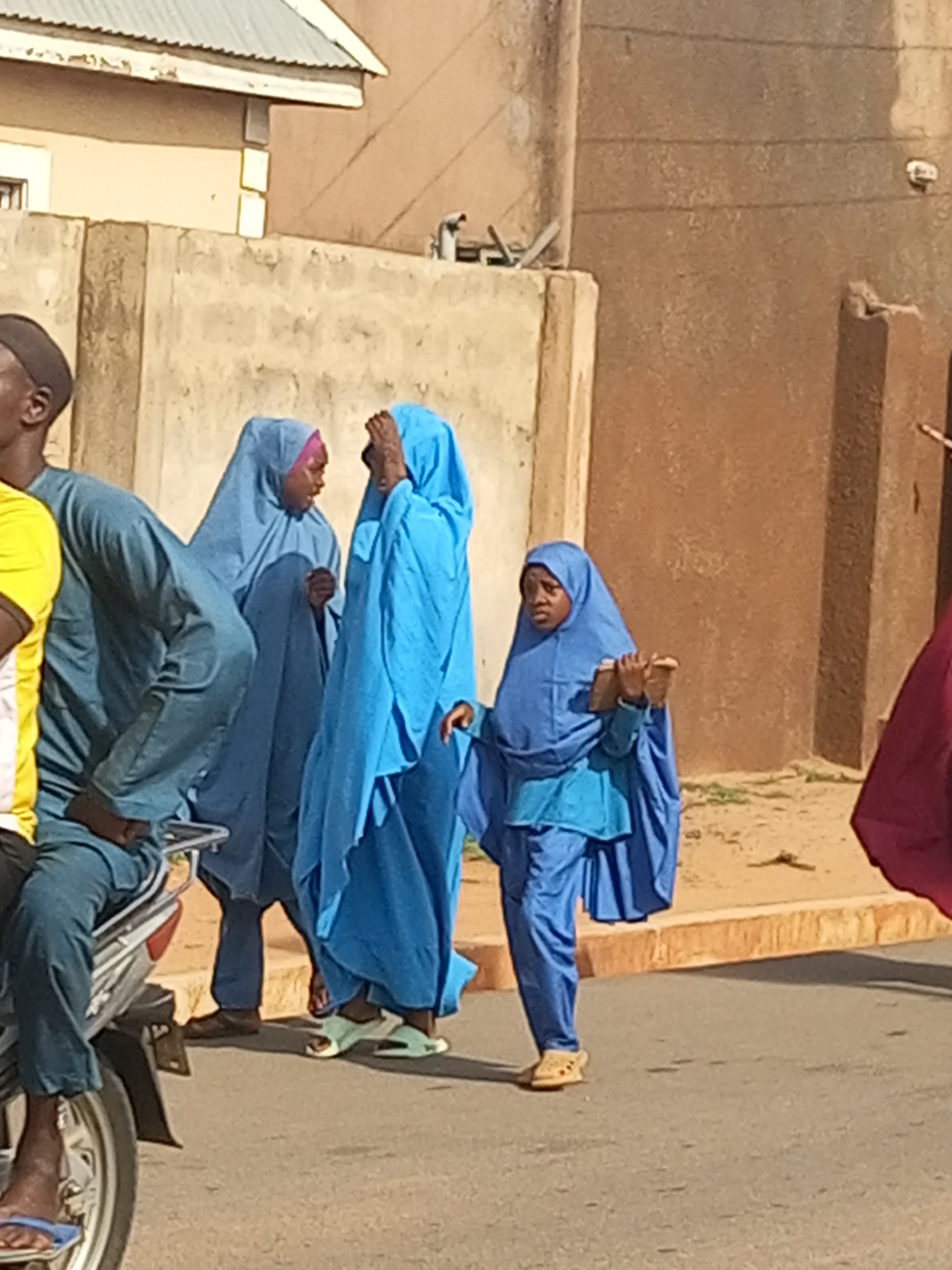 The image depicts a street scene in Nigeria, possibly in the city of Tempure based on the prompt. Several people are present, primarily women wearing blue, full-body coverings or hijabs. One woman is holding a book or tablet. A man is seated on a motorbike, wearing a blue outfit, but with a yellow shirt visible underneath. Other individuals are partially visible on the edges of the frame, one in a red garment. They are standing on or near a paved road next to buildings and walls.