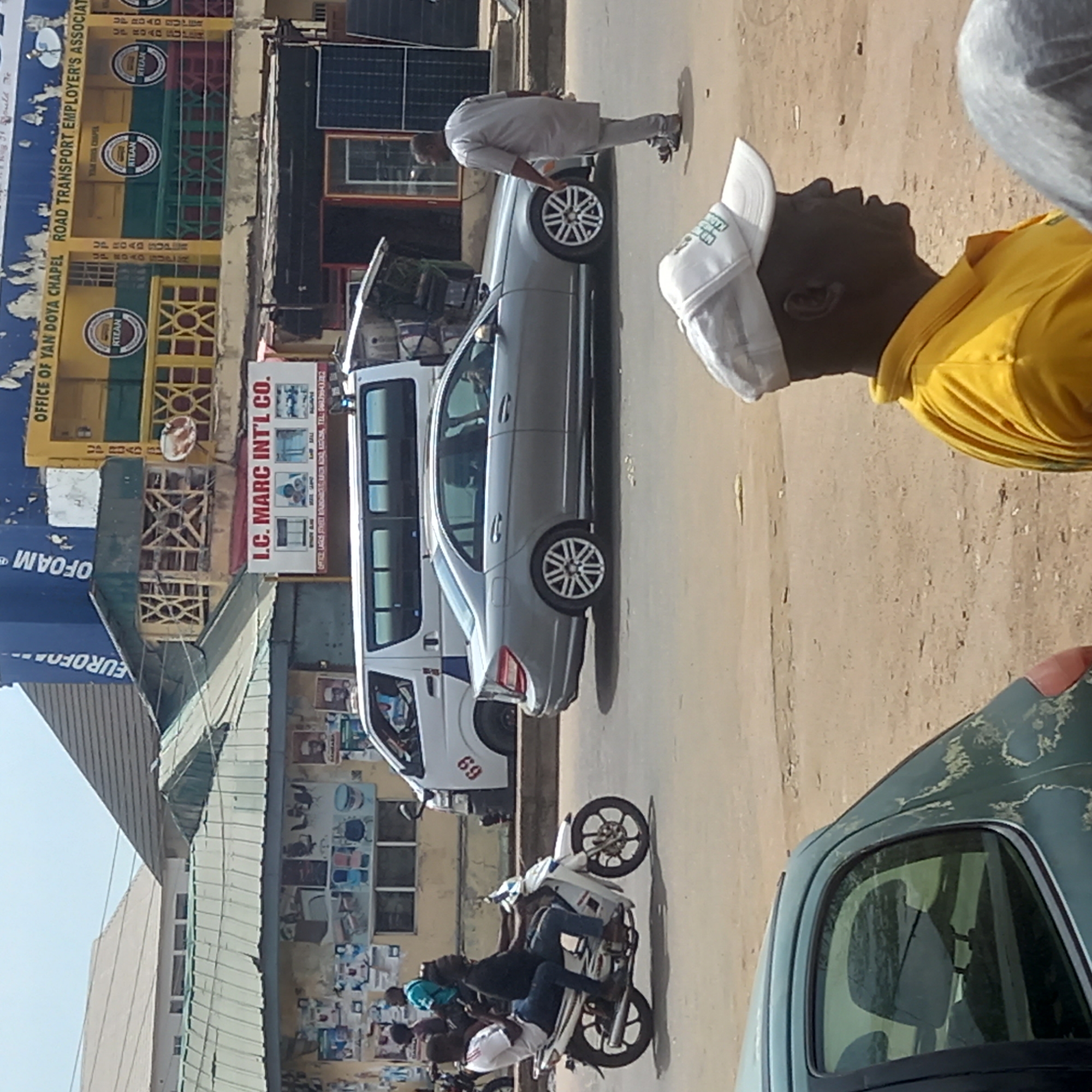 The image captures a bustling street scene in Dadi Riba, Nigeria. A silver Mercedes-Benz sedan is parked parallel to a white minibus with the number "69" on its side. A man in a gray traditional outfit examines the car's rear tire. Another man, wearing a yellow shirt and a white baseball cap, is positioned towards the right, possibly observing the situation. A motorcycle with multiple occupants is positioned in the street.
In the background, buildings display signage, including "OFFICE OF YAN DOYA CHAPEL ROAD TRANSPORT EMPLOYER'S ASSOCIAT," "L.C. MARC INT'L CO.," and "EUROFOAM." Posters and advertisements adorn the walls of the buildings along the street. The road appears to be paved. A fragment of an older vehicle is in the foreground.