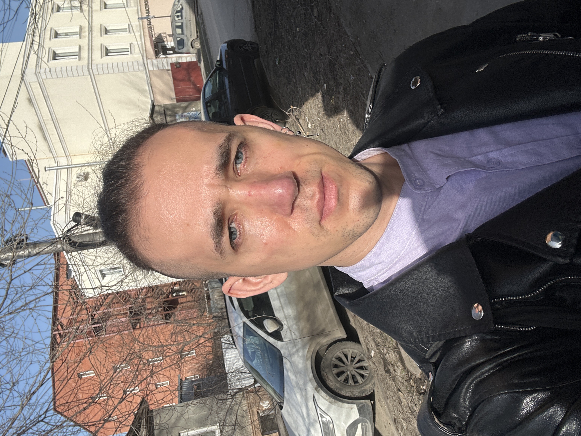 A man takes a selfie outdoors. He wears a black leather jacket and a light purple shirt. A residential street scene is visible in the background, with cars parked on the side of the road. Buildings with windows are also visible. The scene is located in Saratov, Russia.