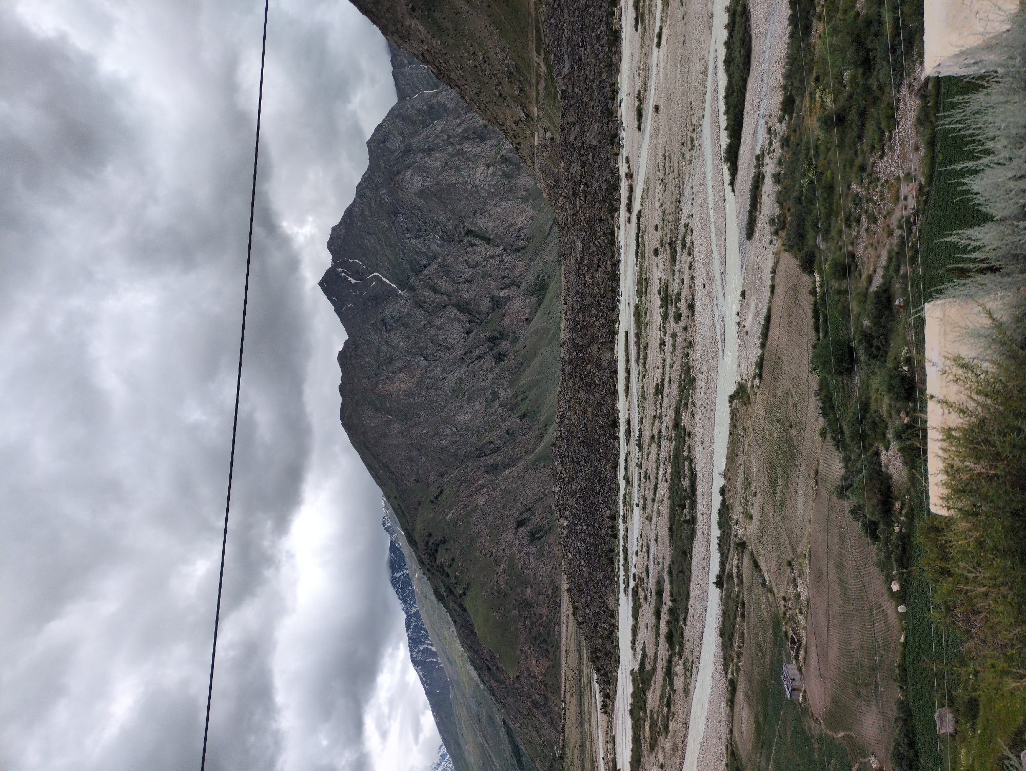 This is a vertical, eye-level shot of a mountainous landscape in Darcha, India, under a heavily overcast sky. A thin, dark power line stretches across the upper portion of the frame, bisecting the cloudy sky. The middle ground is dominated by a wide, rocky riverbed with several channels of pale, flowing water. To the left of the riverbed, in the foreground, is a small, dilapidated building with a corrugated metal roof, situated in a cultivated field. The field itself appears to be plowed in rows. Beyond the riverbed, steep, rocky mountains rise, with patches of green vegetation on their lower slopes. The peaks of these mountains are dark and jagged, with some snow visible at higher elevations. The overall mood is somber and rugged, characteristic of a remote mountain region.