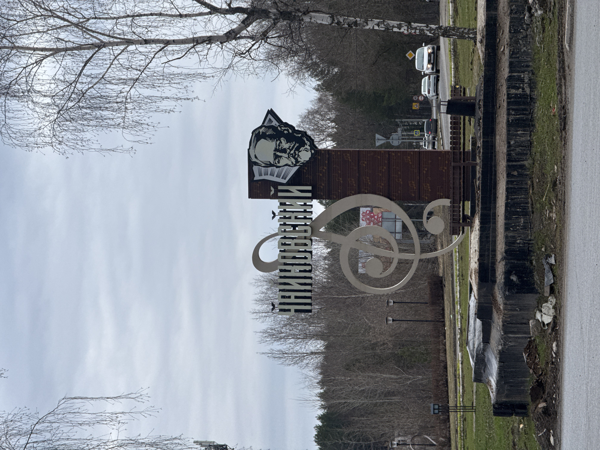The image depicts a monument located in the city of Unknown, Unknown. The monument features a profile of a man above a stylized musical note and the name of the city written vertically in Russian. The monument stands on a roadside, bordered by trees and vegetation. Cars can be seen in the background on the road. The sky is overcast.