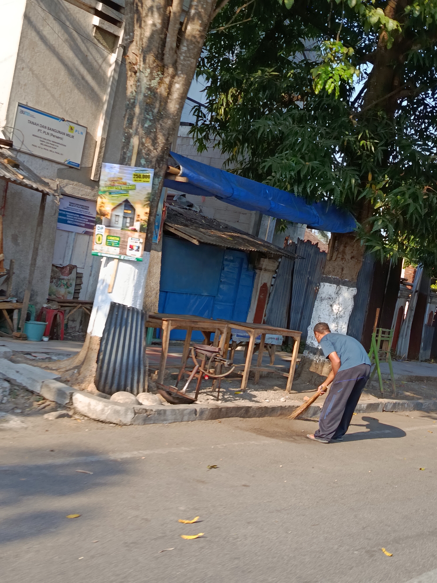 The image depicts a man sweeping the sidewalk in front of a building with a corrugated metal facade. He is wearing a blue t-shirt and dark pants. A large tree with dense green foliage stands to his right, partially shading the scene. In the background, there are several structures with blue doors and corrugated metal roofing. A sign for "PLN TANAH DAN BANGUNAN MILIK PT PLN (PERSERO)" is visible on the wall of the building. The ground is paved with asphalt, and several fallen leaves are scattered across it. The lighting suggests it is daytime, with sunlight casting shadows. The location is specified as Kotakulon, Indonesia.