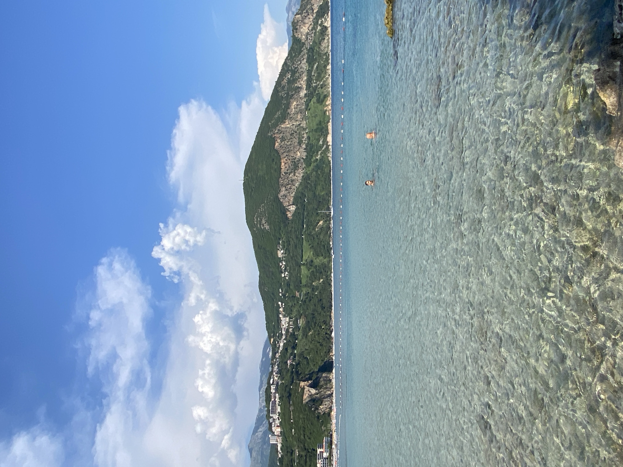 The image depicts a scenic view of the coastline near Prijevor, Montenegro. The foreground is dominated by the crystal-clear turquoise water, revealing the rocky seabed beneath. Two individuals are visible swimming in the sea. In the mid-ground, a marker buoy line separates the swimming area from the deeper water. The background features a lush green mountain slope dotted with buildings, suggesting a coastal town. The sky is a vibrant blue, adorned with fluffy white clouds. The overall scene conveys a peaceful and idyllic summer day at the beach.