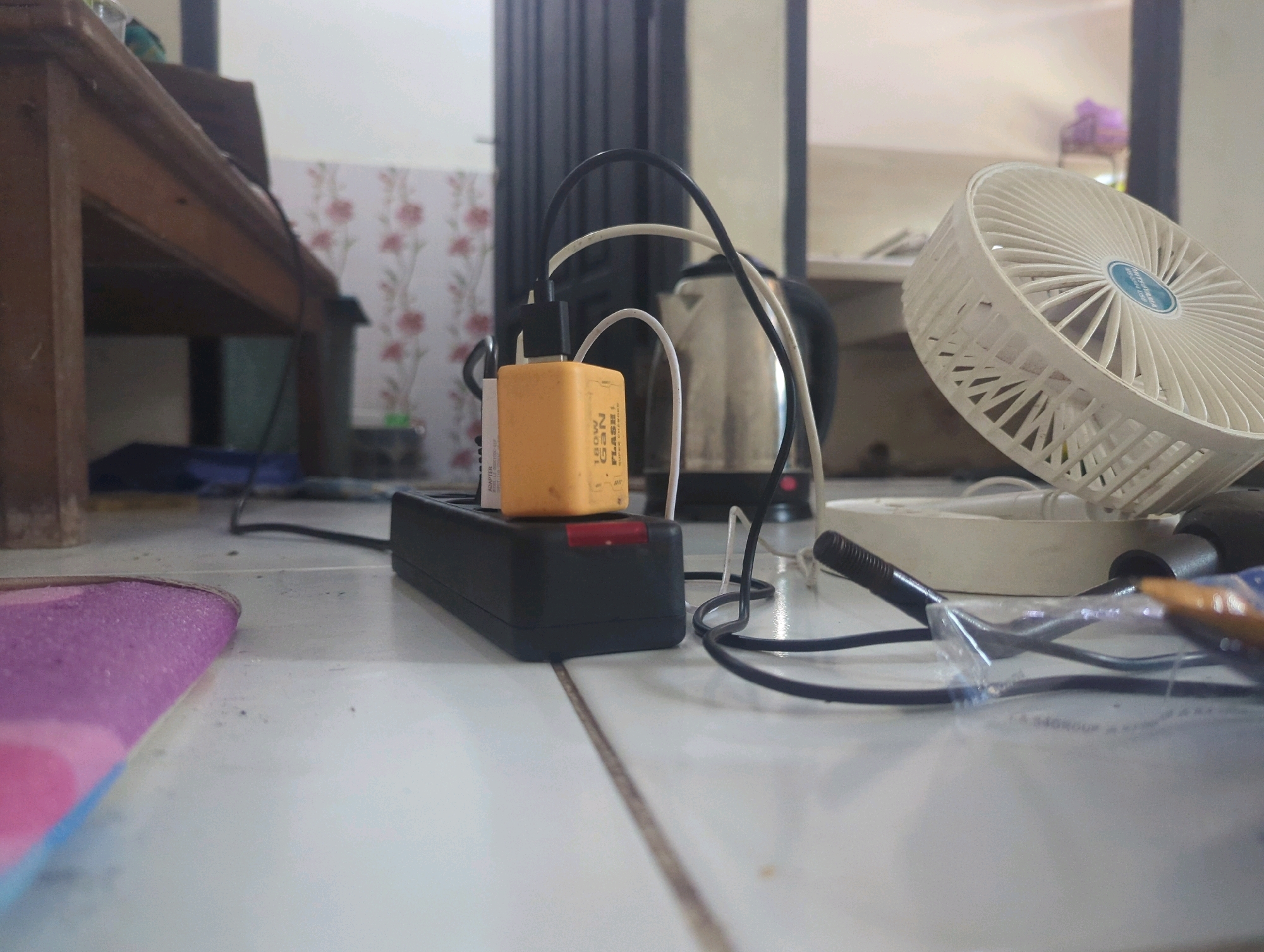 This image shows an indoor scene, likely a room in Batu Belaman, Indonesia. The focus is on a power strip with a yellow charger plugged into it, connected to multiple cables. In the background, a metal electric kettle is visible, as well as a white fan. A portion of a wooden table is also present in the left side of the frame. The floor is tiled and appears somewhat dusty. The overall impression is of a functional, perhaps cluttered, living or working space.