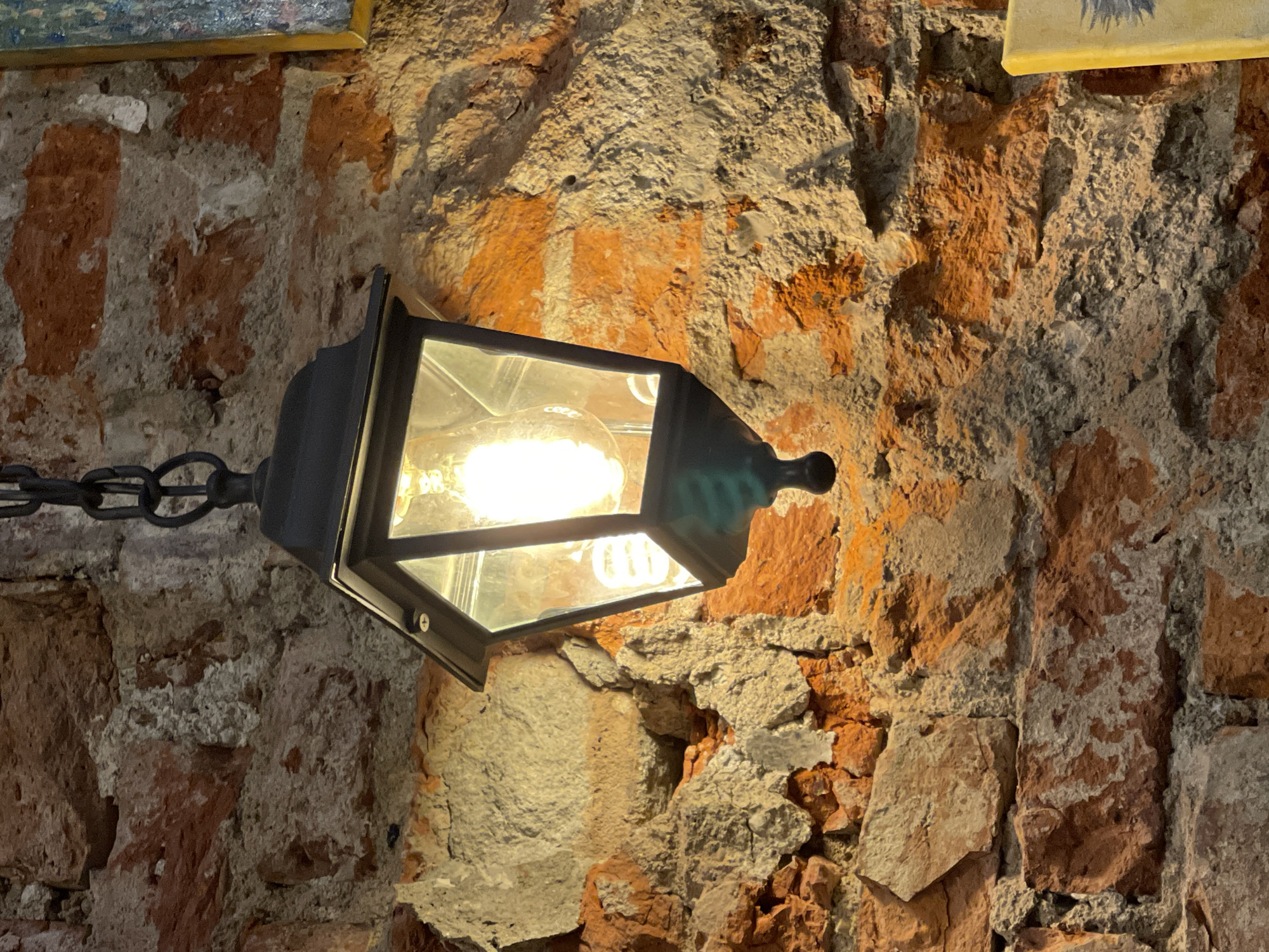 The image displays a vertical, detailed view of an interior brick wall illuminated by a dark metal hanging lantern, located in Kaliningrad, Russia. The wall is composed of irregular reddish-orange bricks, interspersed with thick, rough, light grey mortar and extensive patches of crumbling, off-white plaster or render, indicating significant age and wear.
A black or dark-colored metal lantern-style light fixture is centrally positioned in the upper half of the frame, suspended by a heavy metal chain partially visible at the top. The lantern has clear glass panels, revealing two distinct light sources: a brightly glowing, vintage-style incandescent filament bulb in the foreground, and a coiled white compact fluorescent lamp (CFL) situated behind it. The emitted light casts warm, strong highlights and shadows, accentuating the wall's rough and uneven texture.
In the upper right corner, a small section of a gold-colored picture frame is visible. A yellow object, possibly a book or another decorative item, appears partially at the bottom right edge. There are no people present, and the scene is static, focused on the architectural details and the light source.
