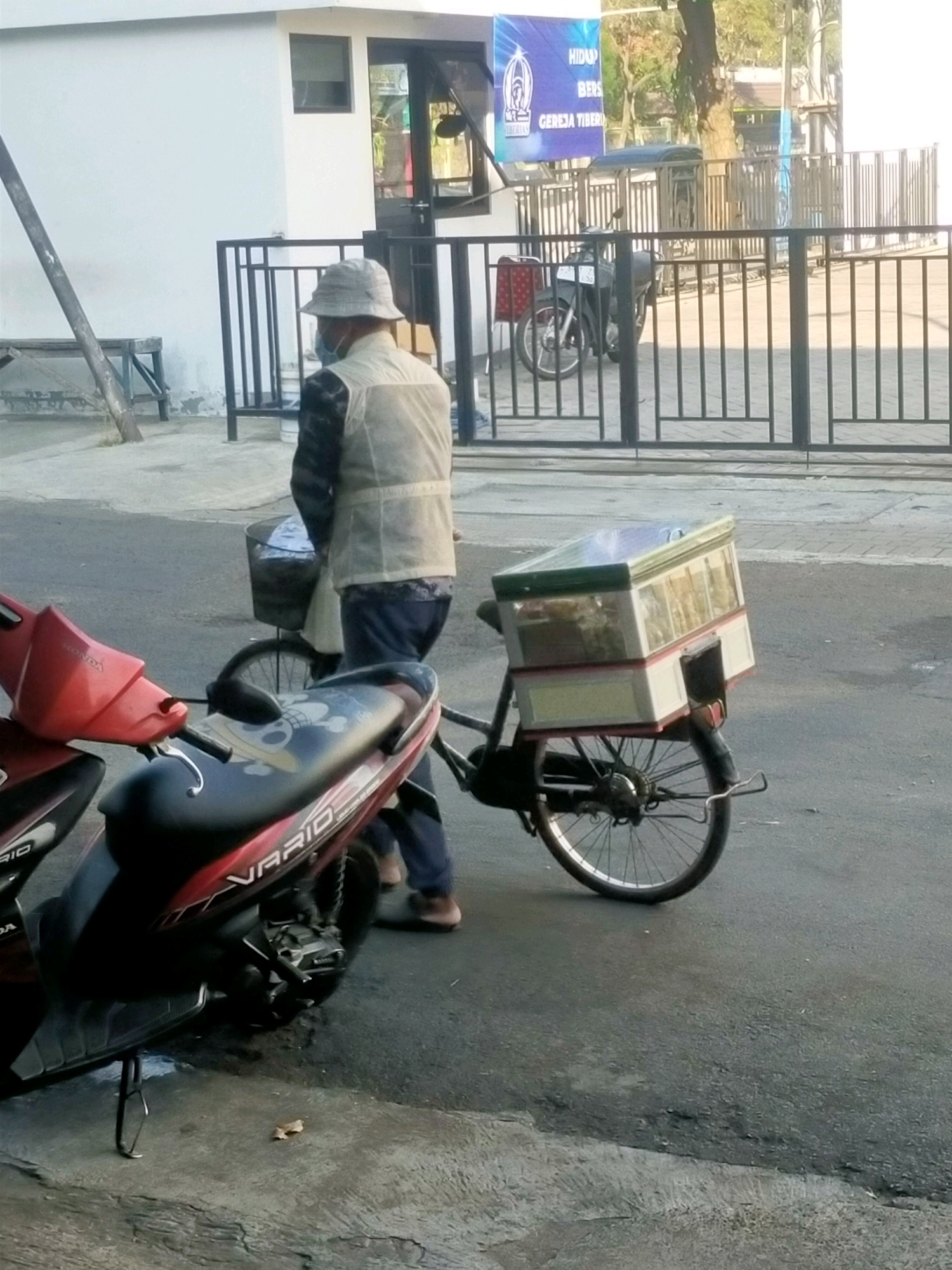 An individual wearing a light-colored vest, a patterned long-sleeved shirt, blue pants, and a bucket hat stands next to a bicycle that has a large white and green cooler box attached to its rear. The cooler box is transparent and appears to contain food items. The bicycle is positioned on an asphalt road, with a red scooter parked in the foreground. Behind the individual and the bicycle, a black metal fence encloses a paved area. To the left of the fence, a white building with a dark-framed window and a closed dark door is visible. A wooden bench is situated near the building. In the background, a banner with the words "HIDUP BERSAMA" and "GEREJA TIBERIAS" is displayed, suggesting the location is near a church in Surabaya, Indonesia. The scene appears to be a street setting, possibly in front of a church or a community building.