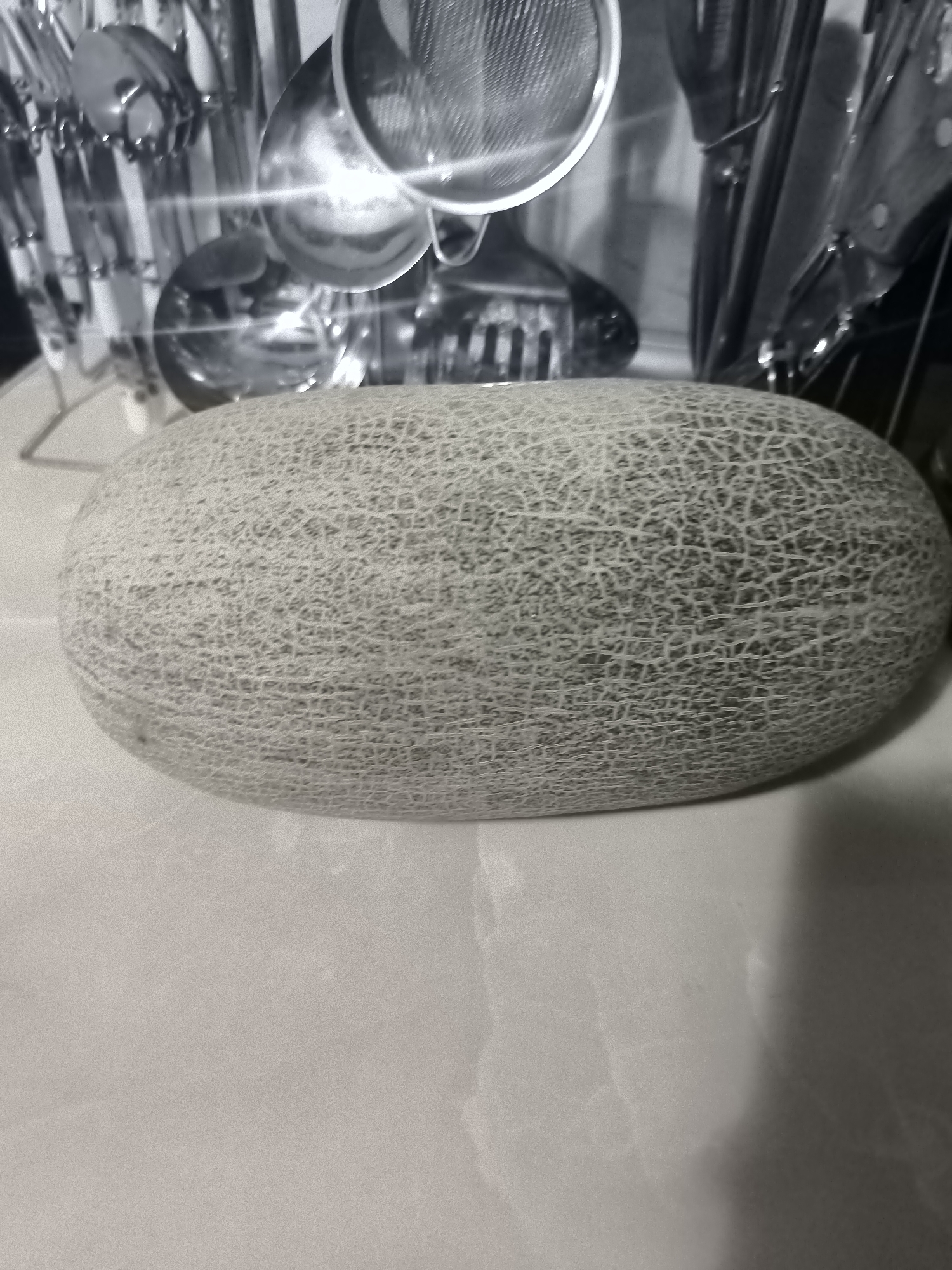 The media file shows an oval-shaped cantaloupe melon resting on a smooth, light-colored surface, possibly a countertop or table. The melon has a textured rind with a net-like pattern of light and dark lines. In the background, out of focus, is a rack holding various kitchen utensils, including ladles and what appears to be a sieve. The overall scene is dimly lit, suggesting an indoor setting, possibly a kitchen. The backdrop behind the utensils has a pattern that resembles tiles or a mural. There is no visible text in the image. The focus is entirely on the melon and the hint of kitchen items in the background, creating a still life composition. The context provided indicates the location is Kokand, Uzbekistan.