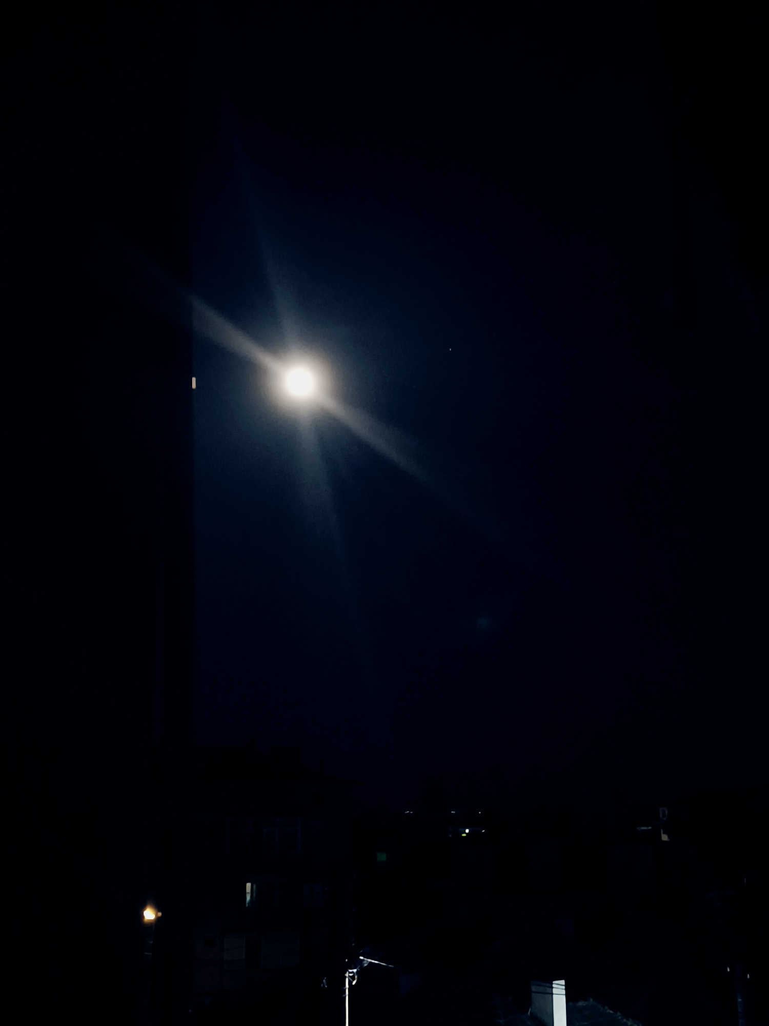The image presents a nighttime cityscape, likely in Svilengrad, Bulgaria. A bright celestial body, possibly the moon, dominates the upper portion of the frame. Its light emanates downwards, partially obscuring the dark sky. The lower section reveals a silhouette of buildings, utility poles, and streetlights. The overall ambiance is one of limited visibility.