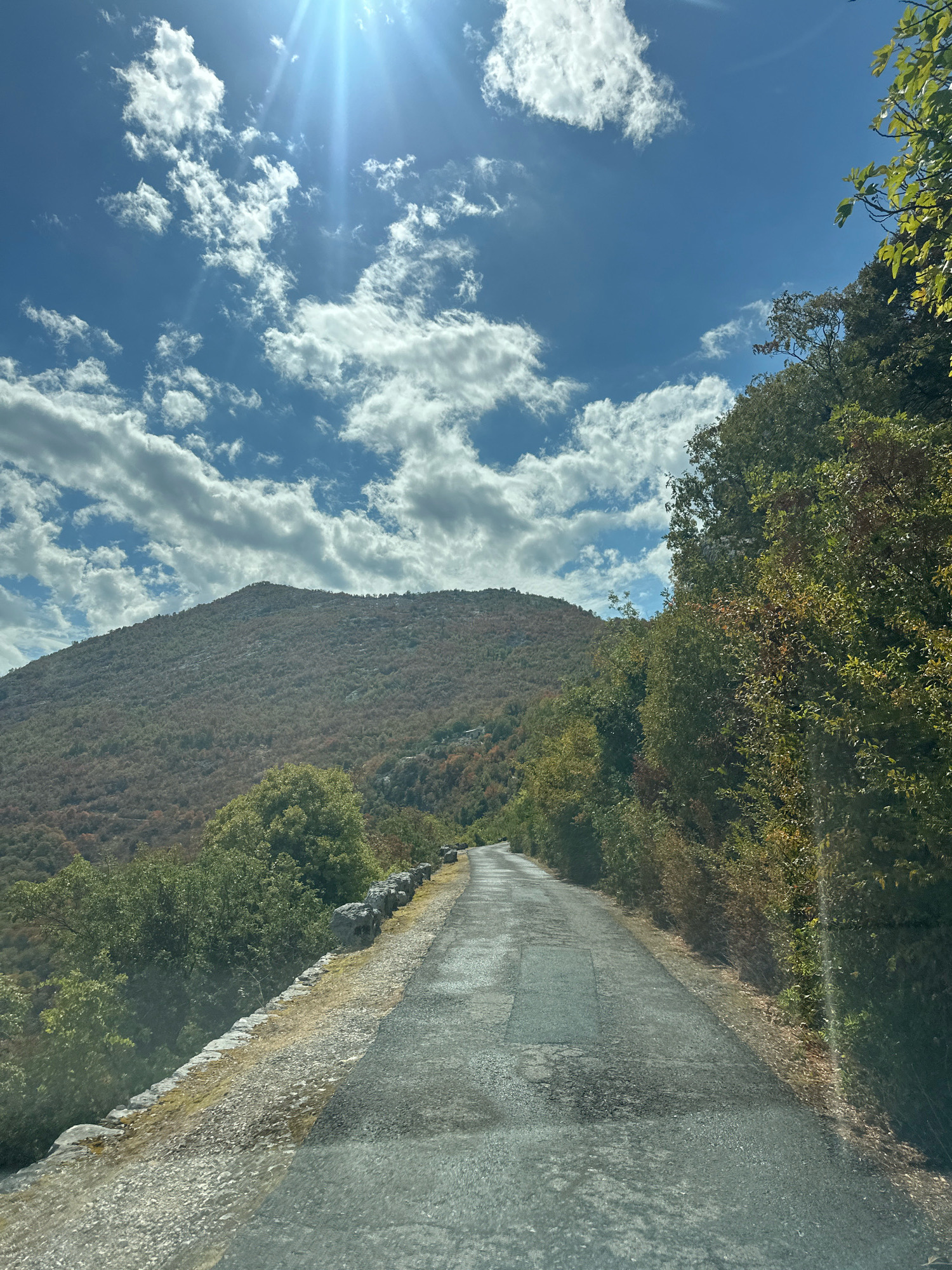The image depicts a narrow, paved road winding through a mountainous, wooded landscape under a bright, partly cloudy sky. The road, which appears to be in fair condition with some patching, curves to the left as it ascends a hill. To the right of the road, dense green foliage forms a wall of trees and bushes, with some patches of autumnal colors visible. A low stone wall lines the left side of the road in the foreground, separating it from a gravelly embankment. In the background, a large, tree-covered mountain dominates the scene, its slopes dotted with varied shades of green and brown vegetation. The sky is a vibrant blue, with scattered cumulus clouds and bright sunbeams streaming down from the upper left corner. The scene is likely captured from a vehicle, as evidenced by the slight glare on the lower part of the image. The location is identified as a road in the vicinity of Cetinje, Montenegro, likely a scenic route in its mountainous surroundings. There are no people or animals visible in the frame.