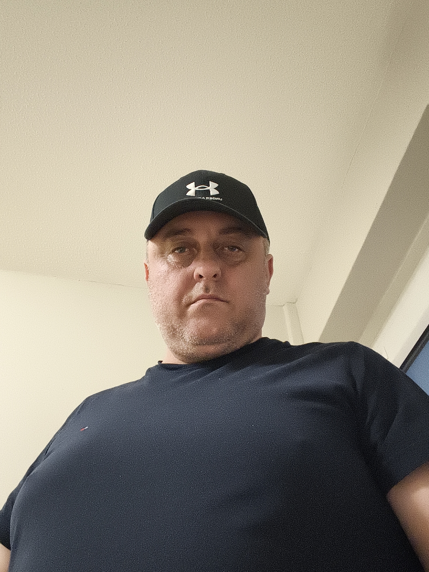 A low-angle portrait captures a middle-aged man looking directly at the camera with a neutral to serious expression. He has short stubble and appears to be Caucasian, with some redness around his eyes. He is wearing a black baseball cap featuring a white Under Armour "H" logo and what appears to be mirrored text "ARBOUR" or similar underneath, possibly indicating a flipped image or reflective lettering. He is dressed in a dark navy or black short-sleeved t-shirt, which has a small, rectangular logo with red, white, and blue horizontal stripes (characteristic of Tommy Hilfiger) on the upper left chest area. The background consists of a plain, light-colored (off-white or cream) ceiling and wall, with a room corner visible on the right, indicating an indoor setting. The lighting suggests artificial indoor illumination. The photograph appears to be a casual portrait taken indoors, within Huntingdonshire, United Kingdom.