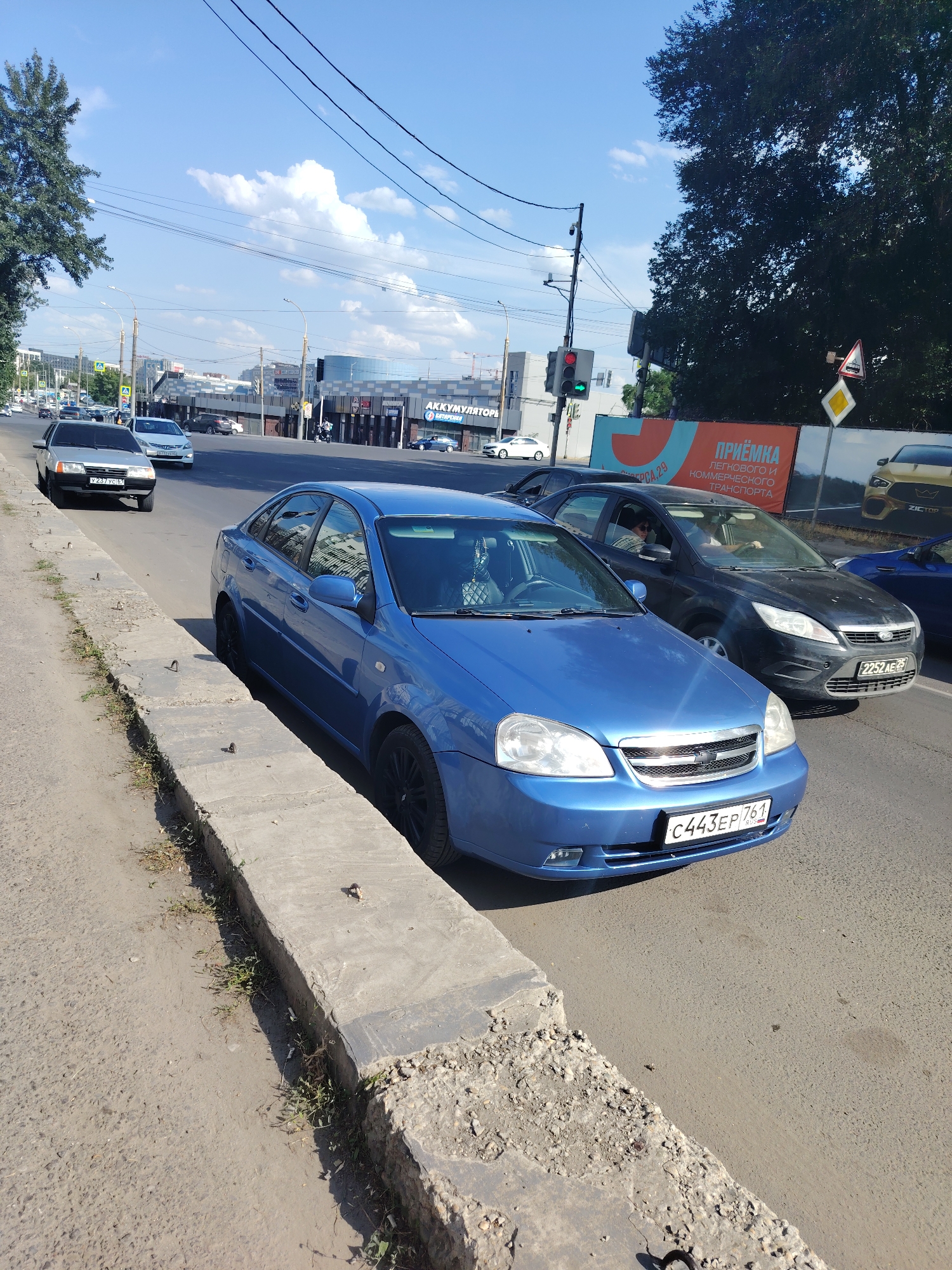 The image shows a street scene in Rostov-on-Don, Russia. In the foreground, a blue sedan is parked along a concrete curb. The car's license plate is visible and reads "C443EP 761 RUS". To the right of the blue car, a dark-colored sedan is in traffic, and its license plate reads "2252AE 76". Further down the street, other vehicles are visible, including a silver sedan with the license plate "Y237VC 76". In the background, commercial buildings and signage are present, including a building with "АККУМУЛЯТОРЫ" (Batteries) written on it. The sky is blue with scattered clouds, and trees line the street. The overall environment suggests a sunny day in a moderately busy urban area.