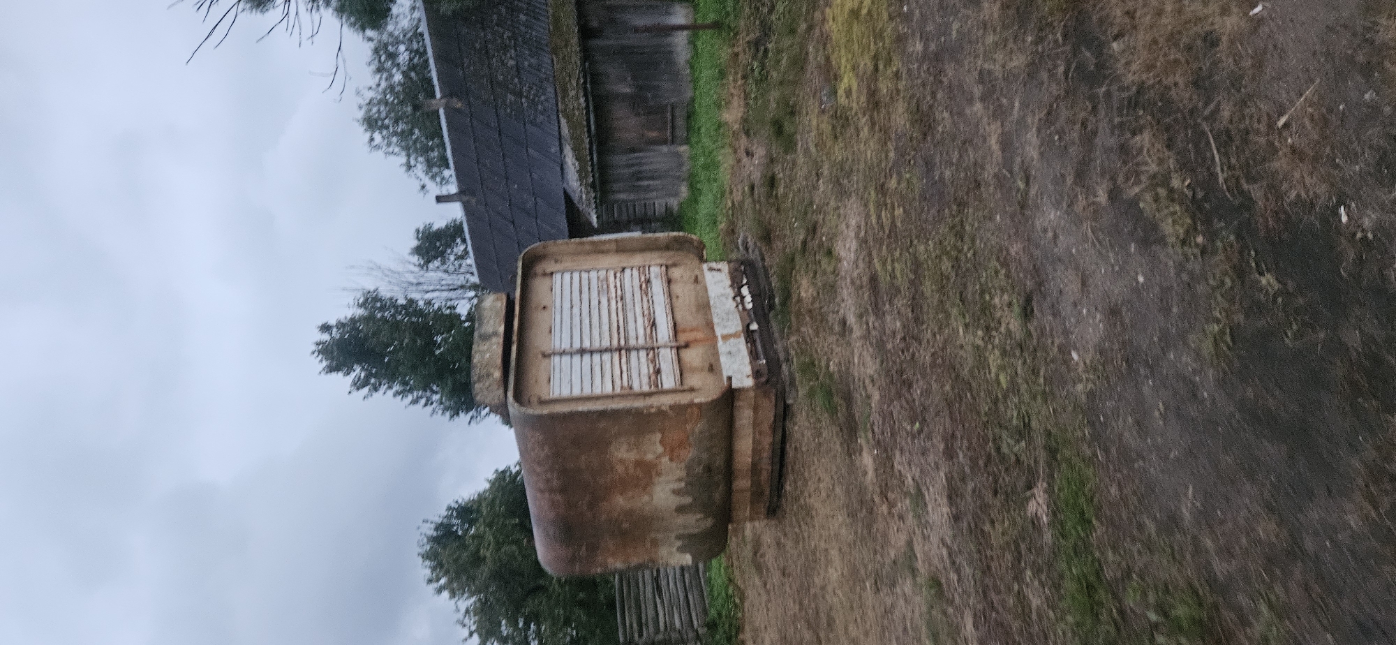 An outdoor scene in Liozna, Belarus, features a large, rusted, metallic object with a slatted opening. The object appears to be a piece of industrial equipment or a storage container, situated in a grassy and dirt area. Behind it, a rustic wooden building with a dark, corrugated roof is visible, along with a wooden fence. Mature trees with green foliage are scattered throughout the background, and the sky is overcast with gray clouds. The ground is uneven, with patches of grass and dirt, suggesting a rural or semi-abandoned setting.