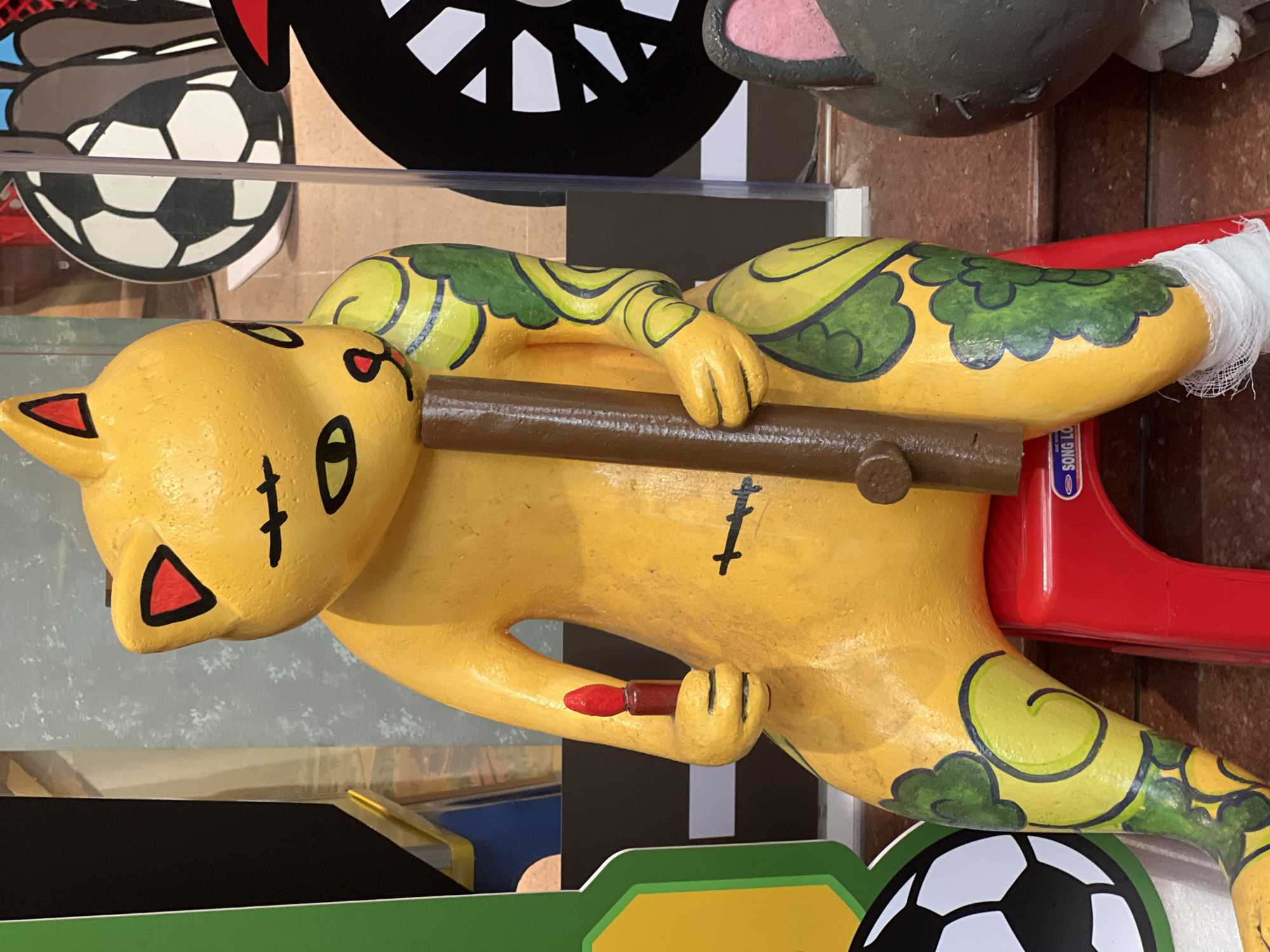 A bright yellow, cartoonish cat statue is seated on a small red stool, holding a brown cylindrical object in its right hand and a red candle in its left. The cat has green, swirling patterns painted on its arms and legs. A white bandage is wrapped around its left ankle. To the left of the cat, a green and yellow cutout of a soccer ball is visible. To the right, a dark grey cat statue with pink ears and black stitching across its body is partially visible. The background appears to be a store or display area, with blurred images of a soccer ball and a motorcycle wheel. The scene is likely located in Hanoi, Vietnam, given the style of the statue and the presence of the brand "SONG LO" on the stool, which is a Vietnamese brand.