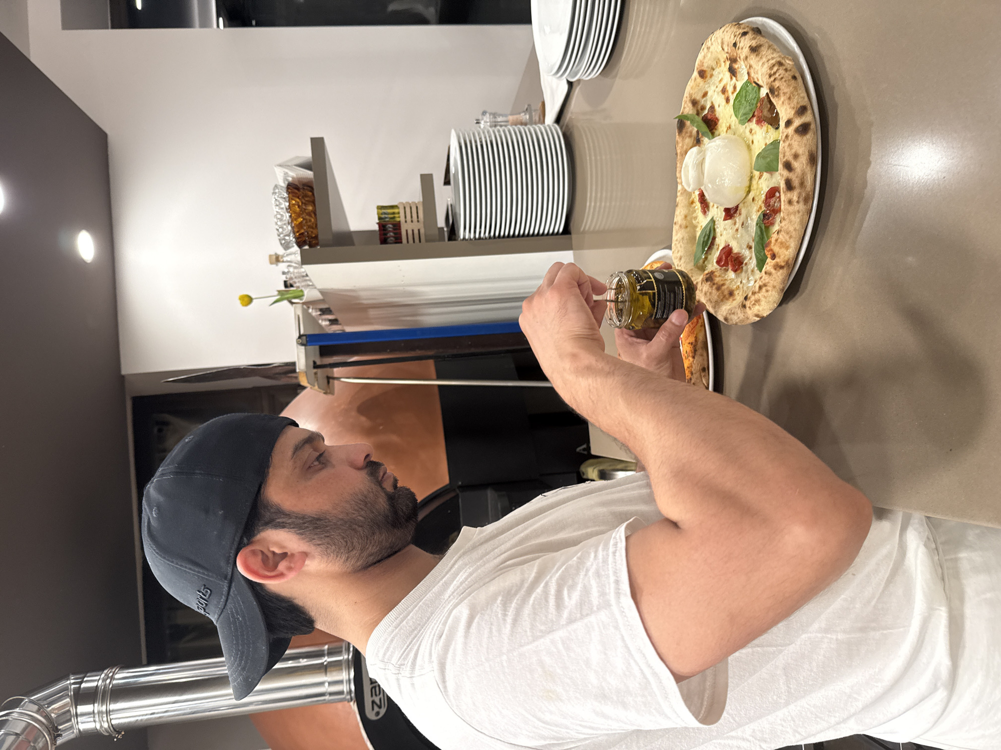 A man wearing a white t-shirt and a black baseball cap is standing at a counter, preparing a pizza. He is holding a small jar of olive oil with a utensil in it. In front of him is a freshly made pizza with burrata cheese, cherry tomatoes, and basil. The background shows a pizza oven and shelves stocked with plates and other items. The image appears to be taken in a restaurant or pizzeria in Trento, Italy.