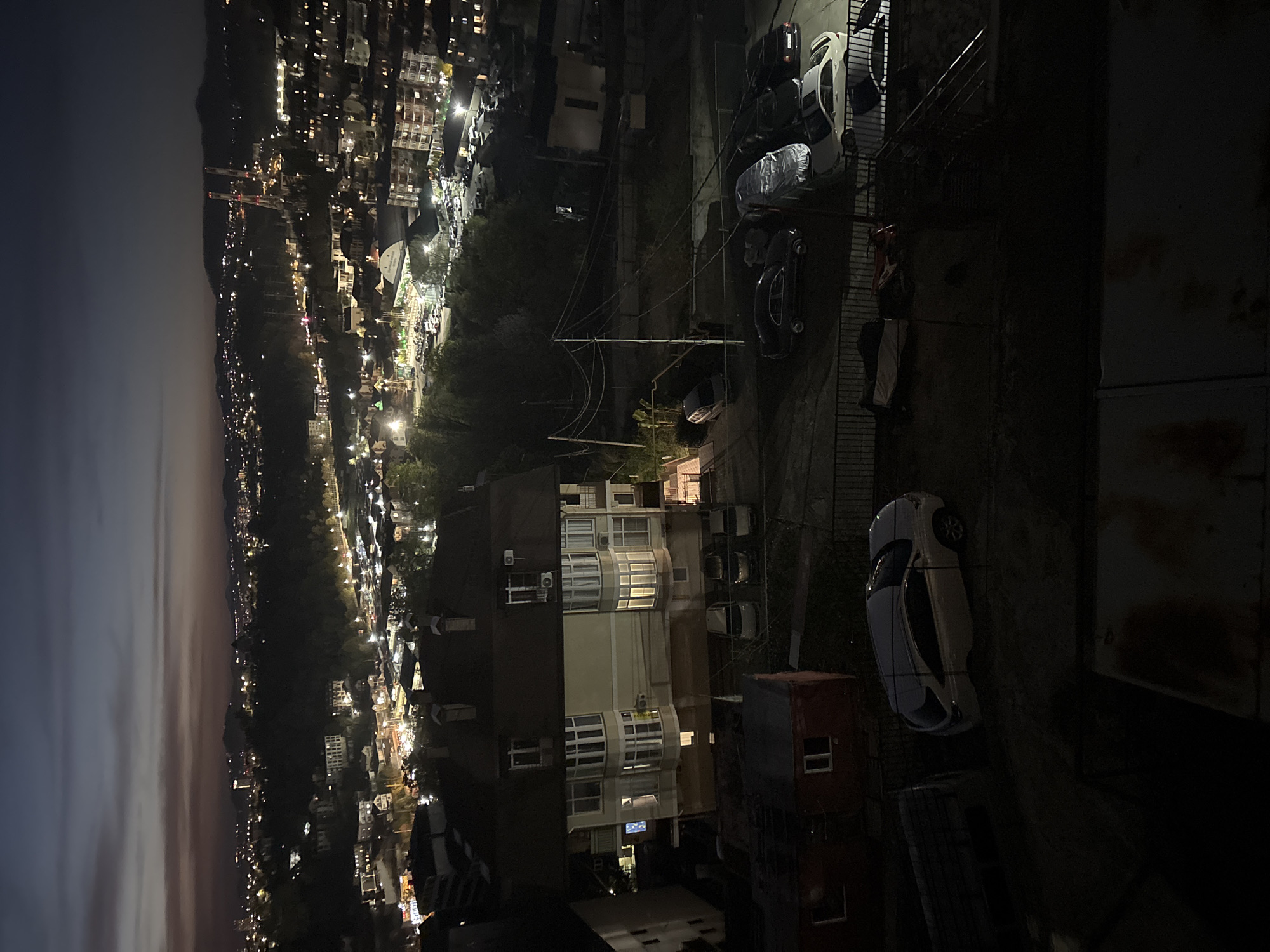 The image shows an aerial view of a residential area in Sochi, Russia, at night. The sky is dark with a hint of twilight. The city extends along a mountain ridge, with numerous lights from buildings and streets illuminating the landscape. In the foreground, a multistory building with lighted windows stands prominently. An alleyway lined with parked cars separates the building from a fence and a narrow pedestrian walkway. A few individuals are visible walking along the walkway.